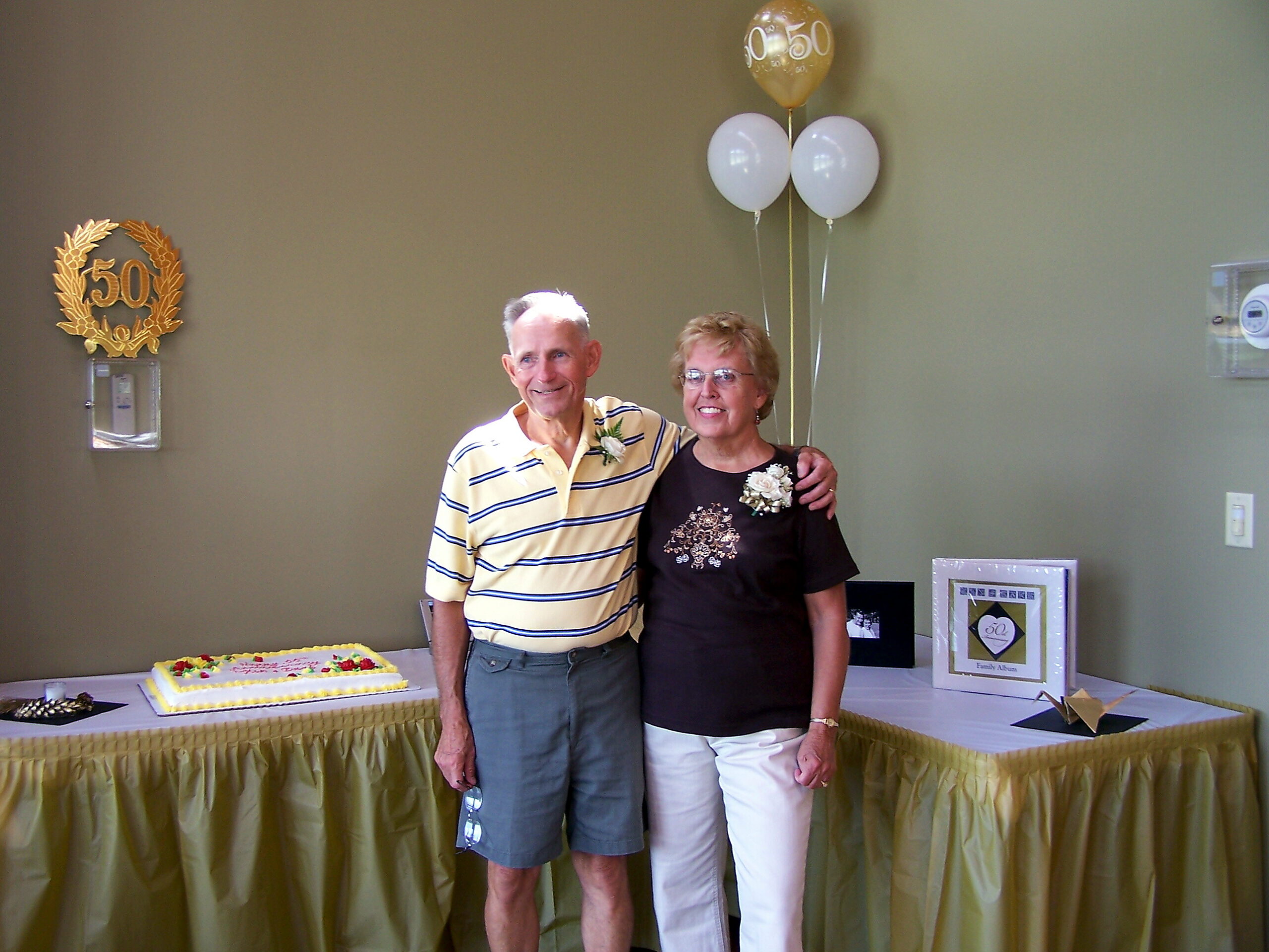 Older couple posing at a 50th anniversary party with balloons and cake behind them.