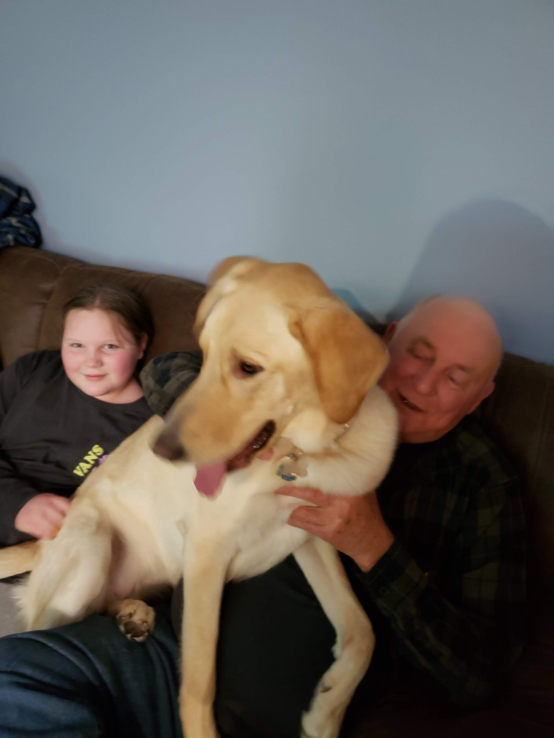 Girl and man sit on a brown sofa with a large light-colored dog between them, tongue out and facing forward.
