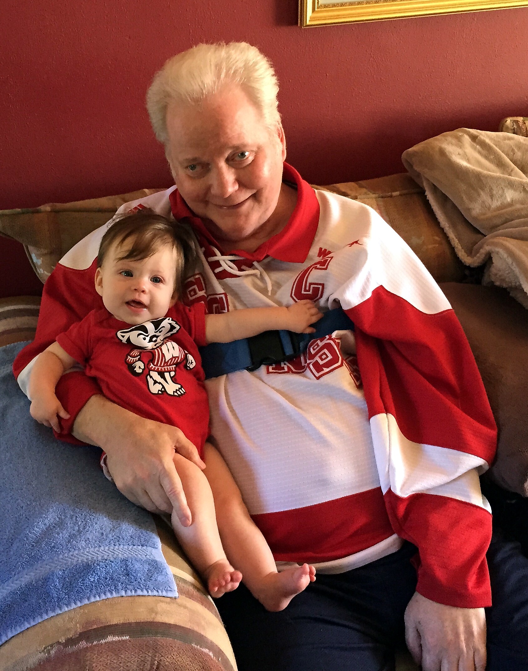 Older man with white hair smiling while holding a baby on a couch in matching red and white hockey jerseys.