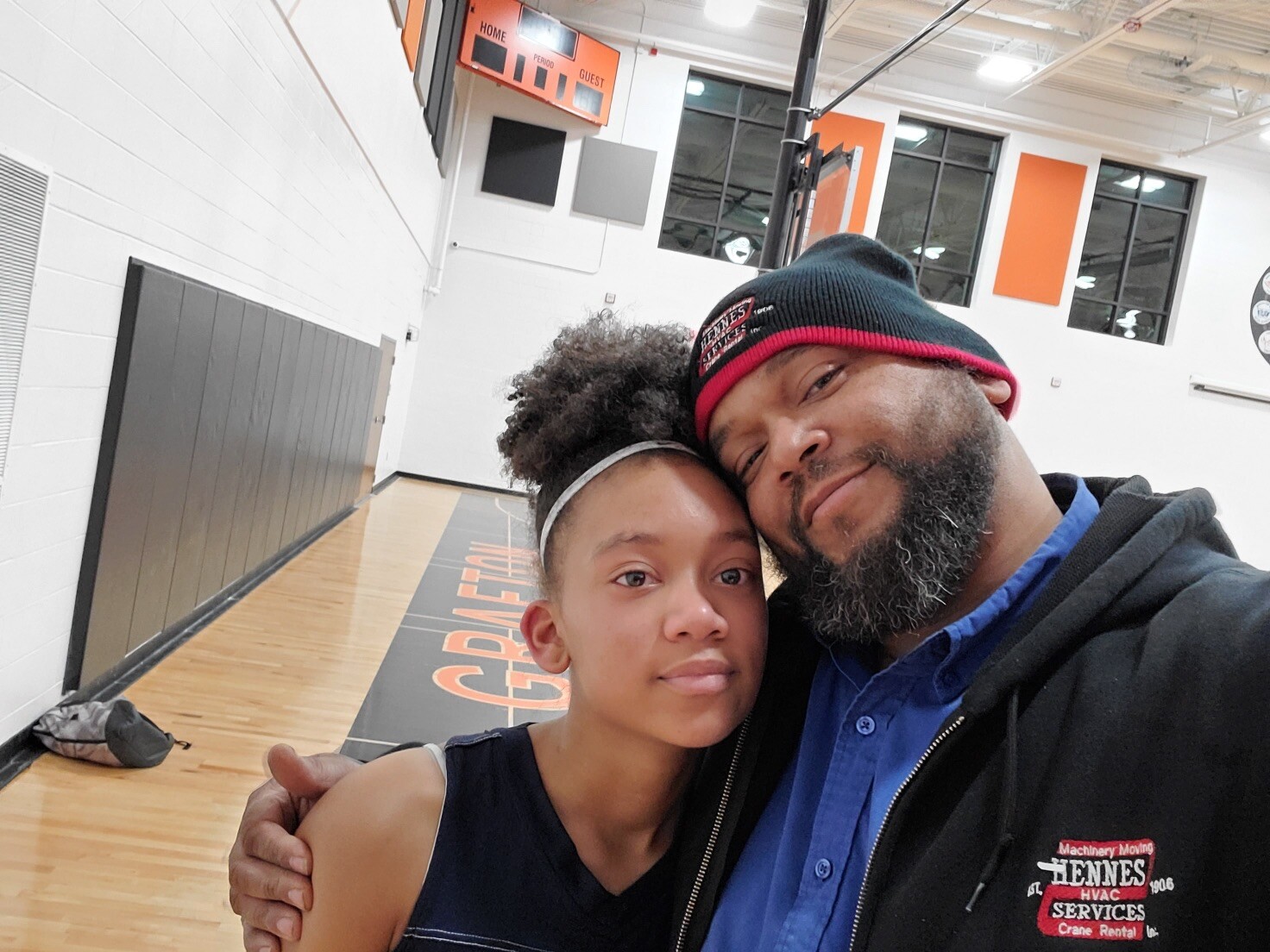 A man and girl take a close-up selfie in a gymnasium, smiling and hugging, with a basketball court in the background.]