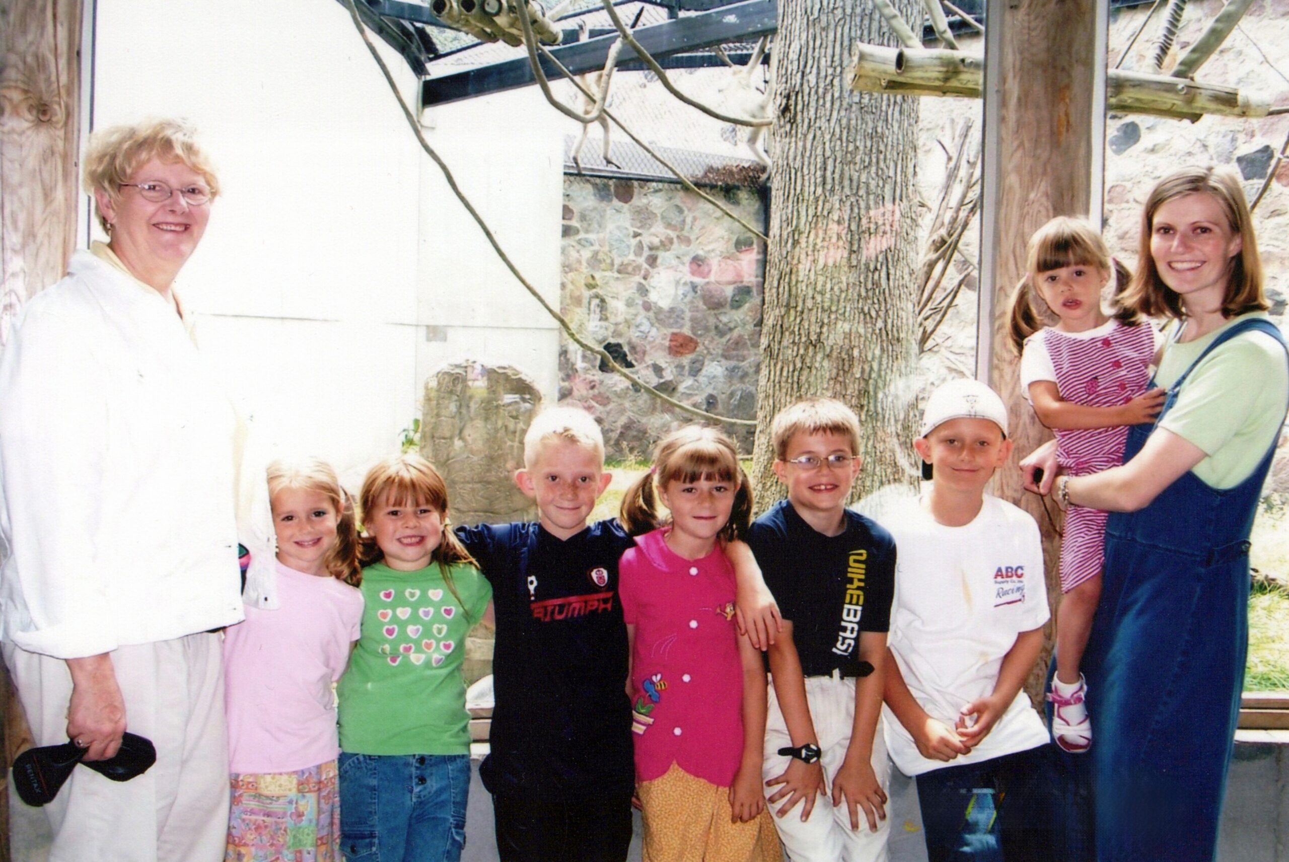 Group of smiling children posing with two adults outdoors near a tree and stone wall; everyone looks at the camera.