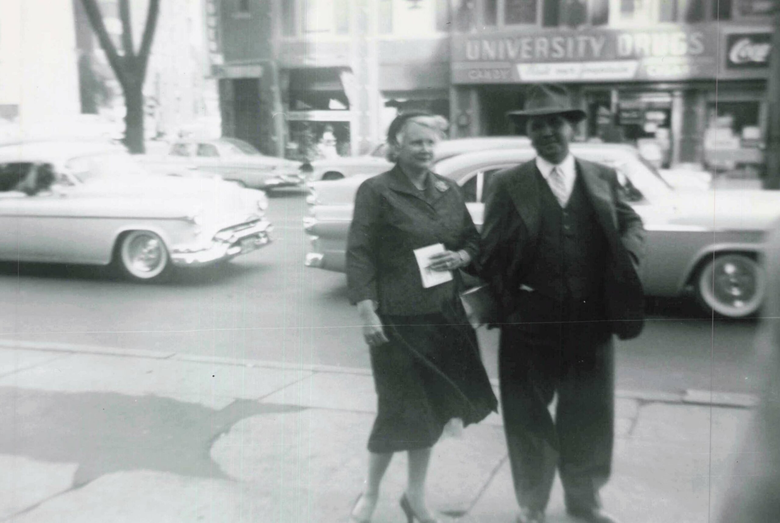 A woman in a dark coat holding papers walks beside a man in a suit and hat on a city sidewalk, vintage cars behind them.
