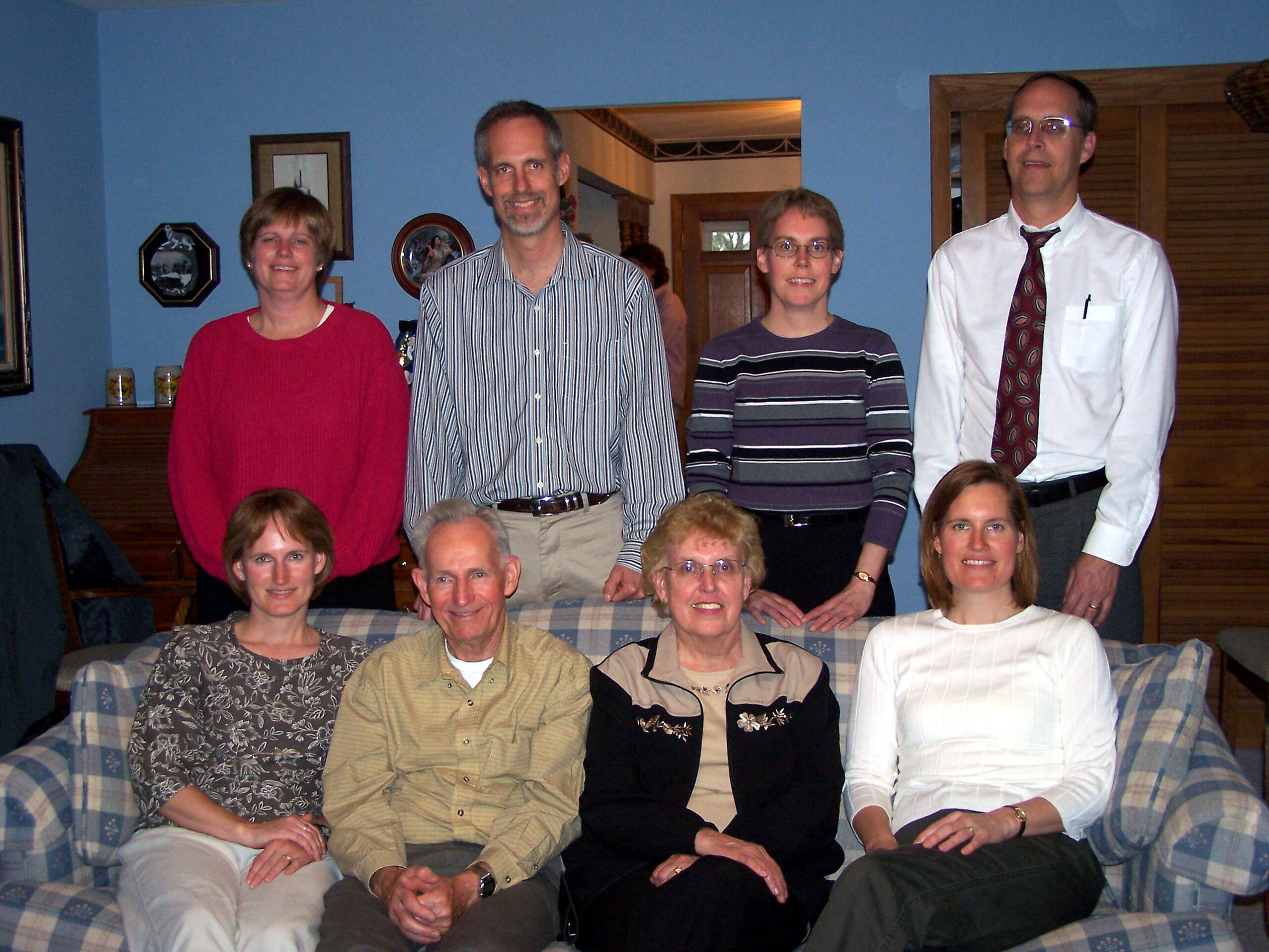 Group of nine adults posing for a family photo in a blue-walled living room, seated on a plaid sofa.