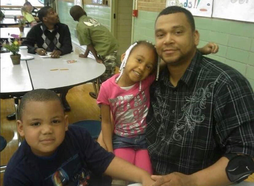 Man with two smiling children posing in a school cafeteria at a round table with a green wall in the background.
