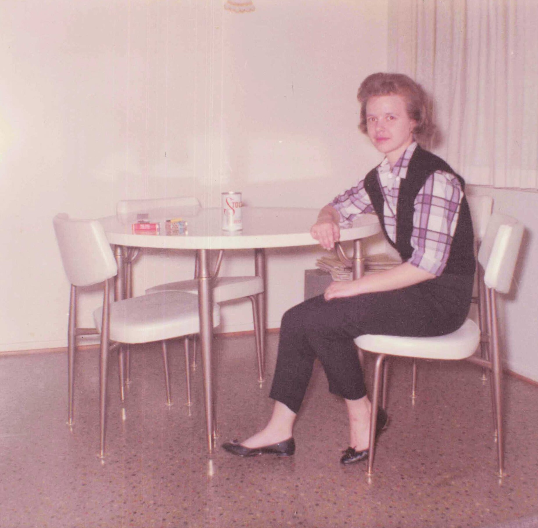Woman in a plaid shirt and dark vest sits at a round table in a beige room; a can is on the table nearby.