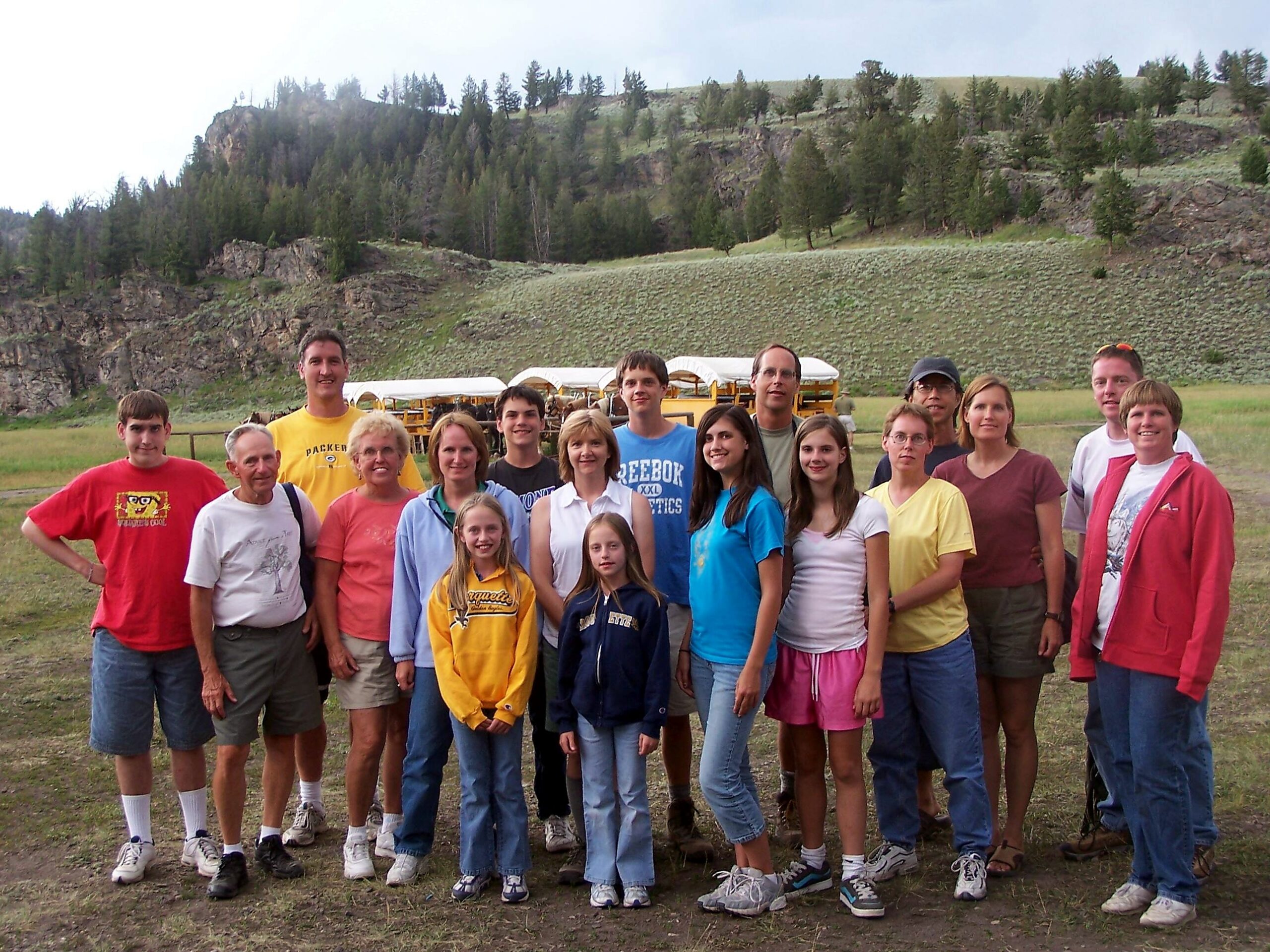 Group of smiling people posing outdoors in a grassy field with a forested hillside in the background behind them.