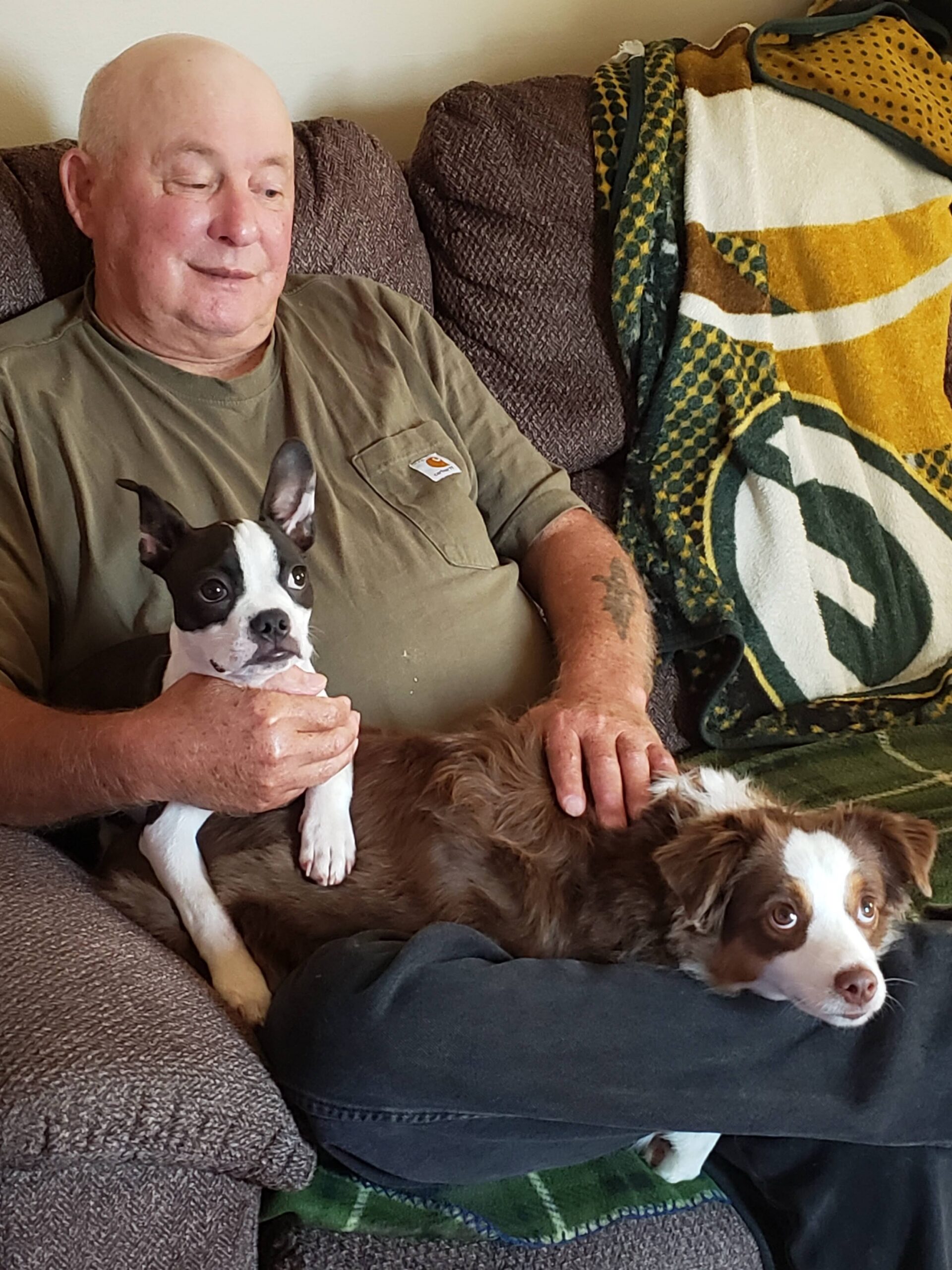 Older man sits on a sofa holding a small black-and-white dog while another brown-and-white dog rests beside him on his legs.