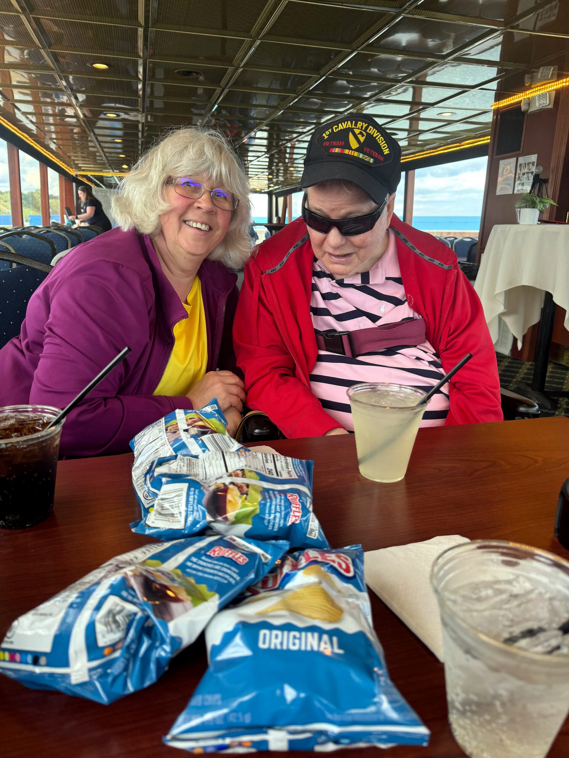 Smiling elderly couple seated at a boat's table with snack bags and drinks in front of them, water visible outside the windows.
