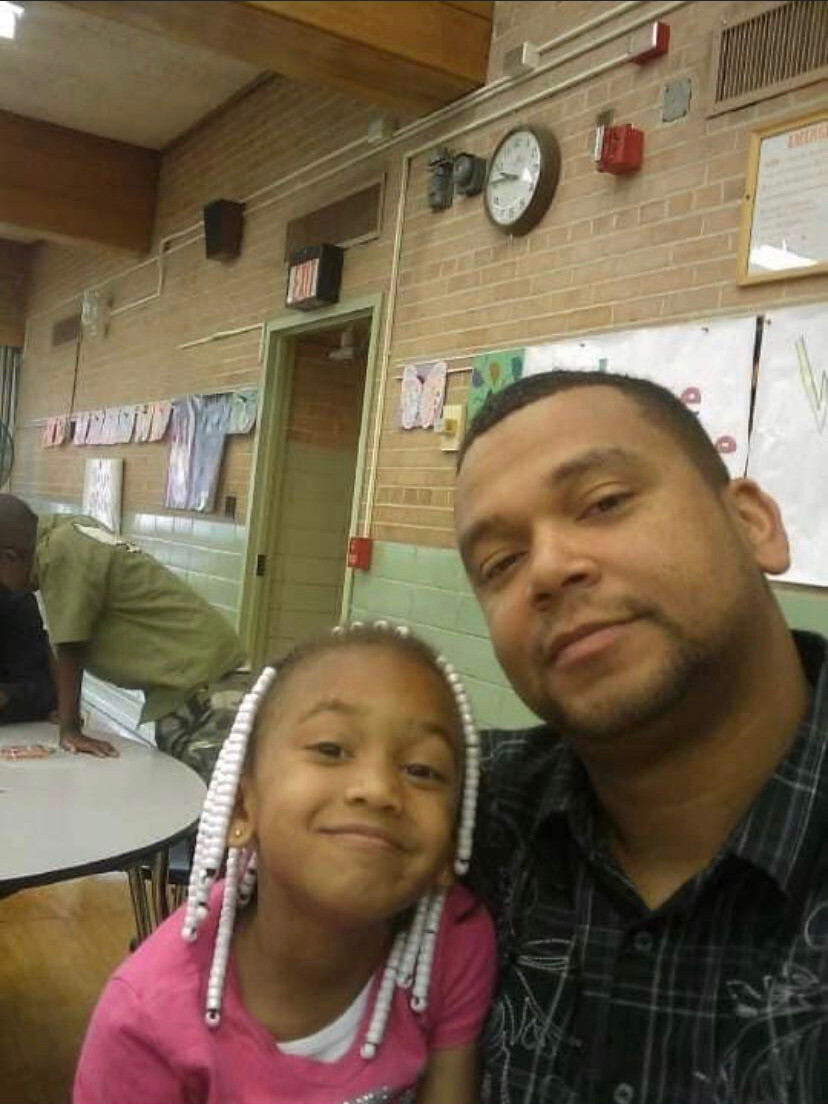 Man and young girl smiling at the camera in a classroom, with brick walls and children’s artwork in the background.
