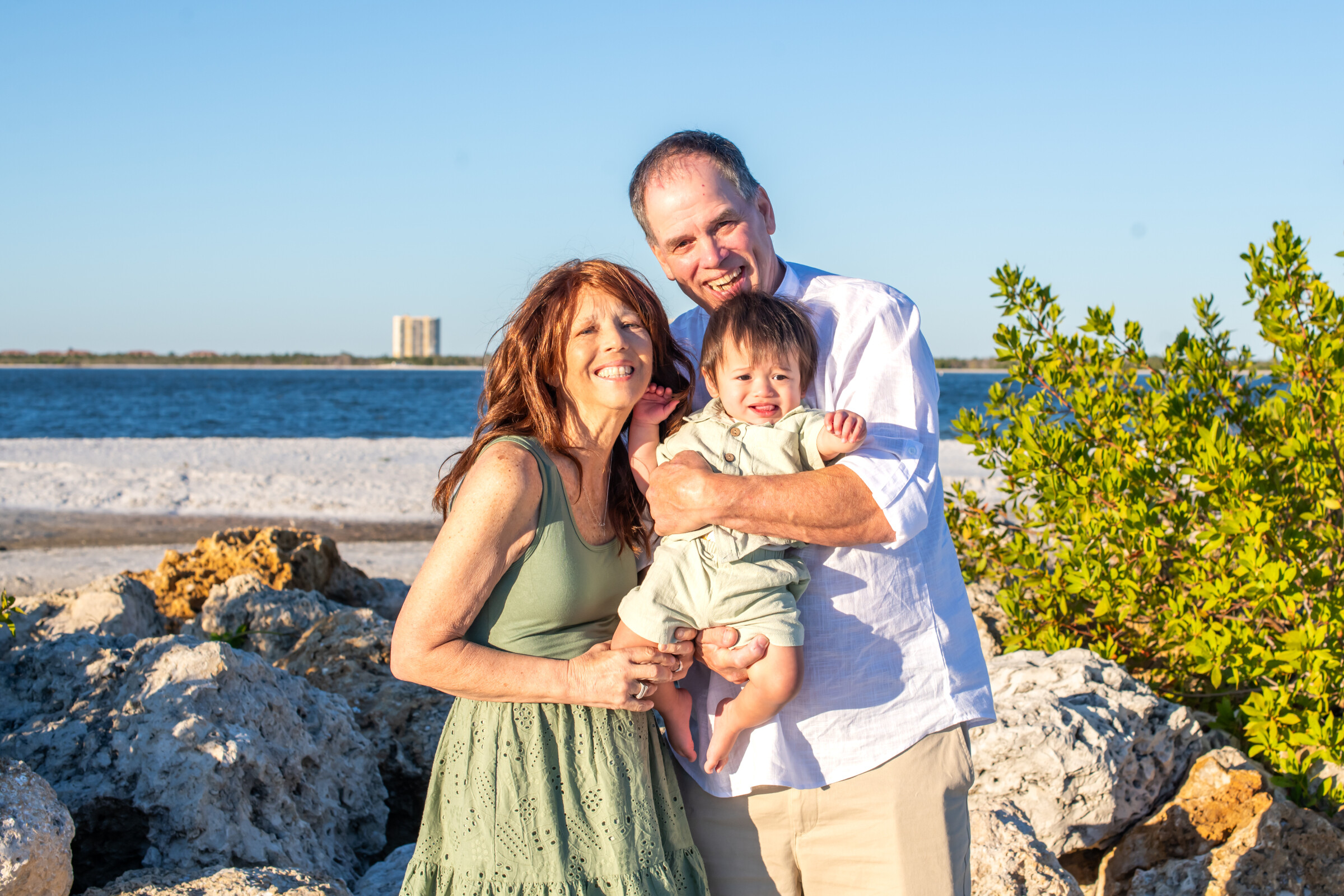 Happy family of three posing on a rocky beach with blue water and clear sky in the background (mother, father, and baby boy).