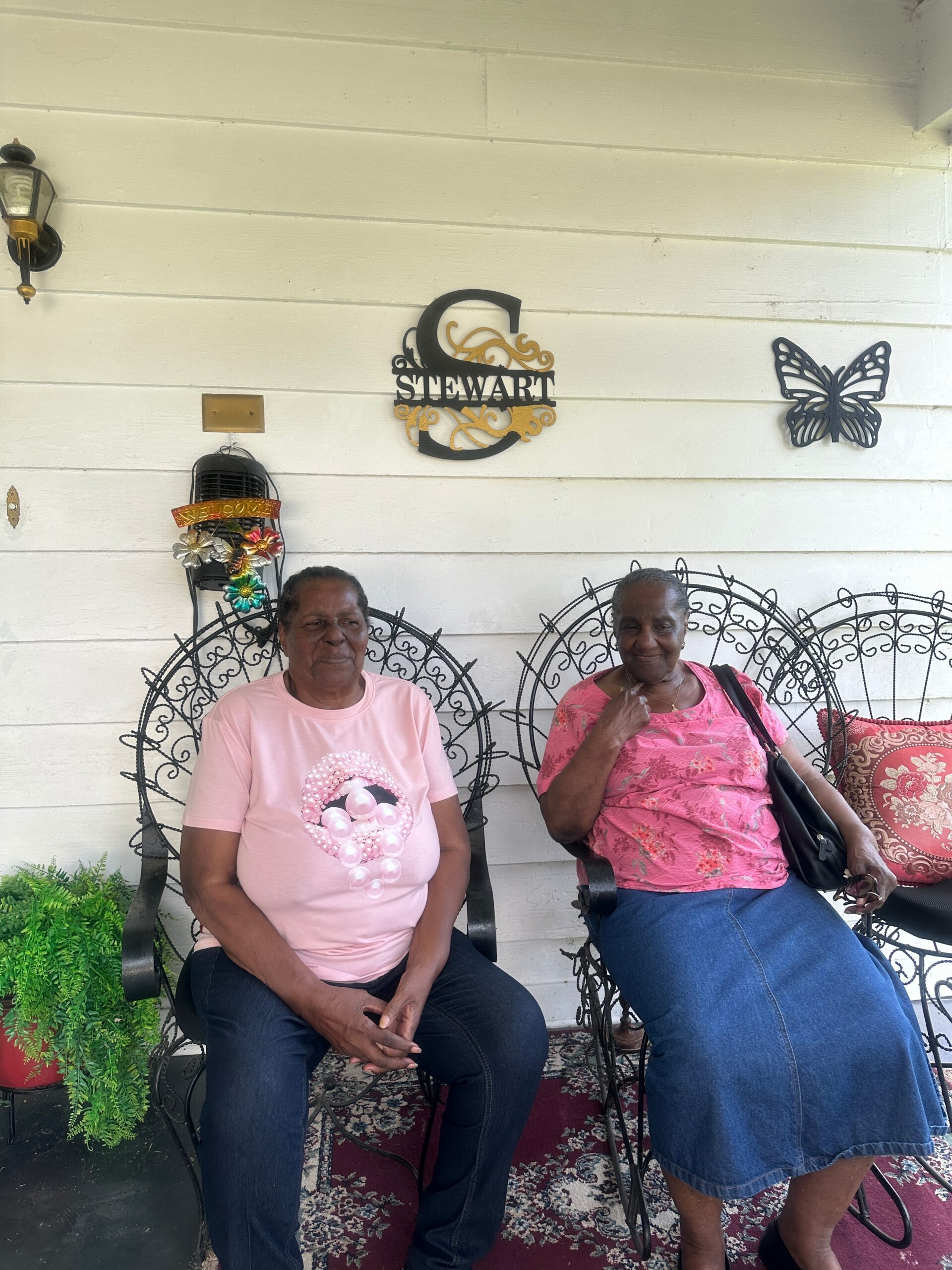 Two elderly women sit on ornate black metal chairs on a porch, with a white wall and a decorative 'Stewart' sign and butterfly wall art behind them.