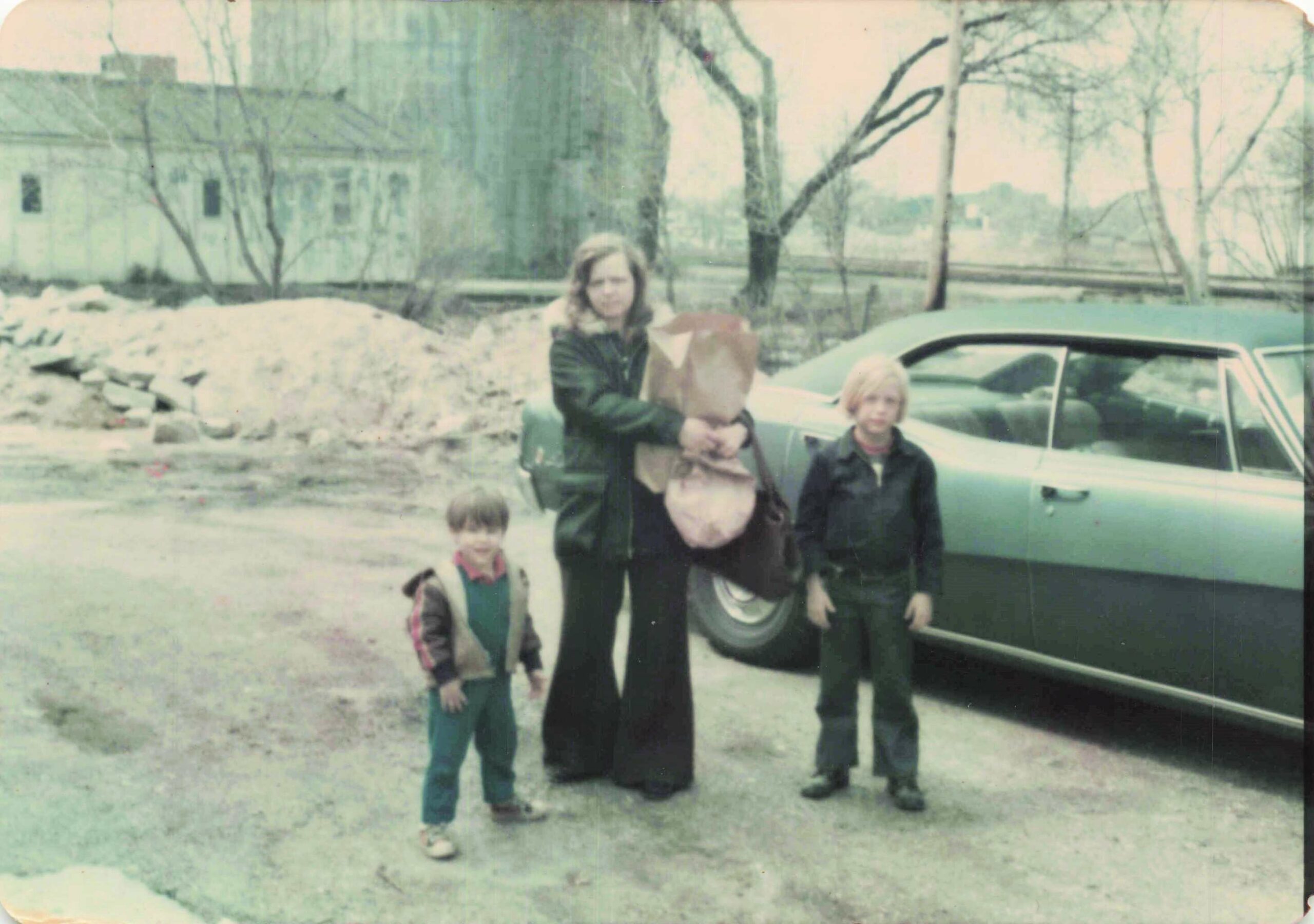 Woman in a dark coat holding a paper bag stands with three children beside a light-colored car in a muddy yard with bare trees and a building in the background.