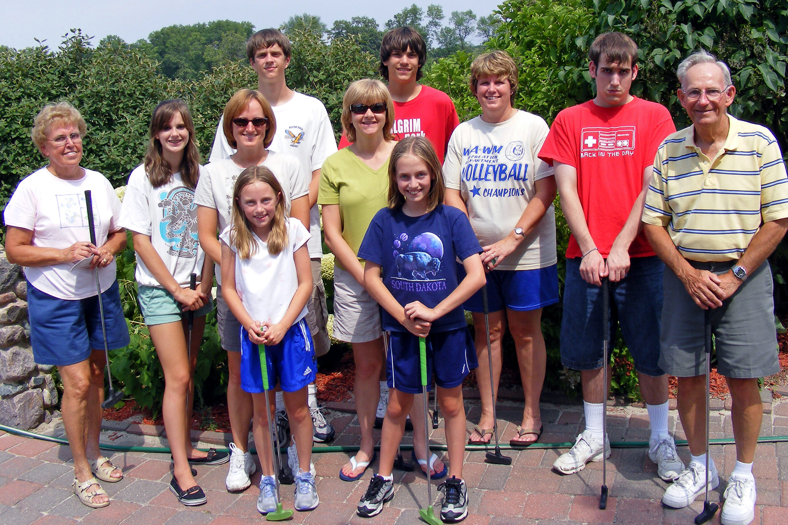 Group of ten people posing outdoors with golf clubs, standing on a brick patio with greenery behind them.