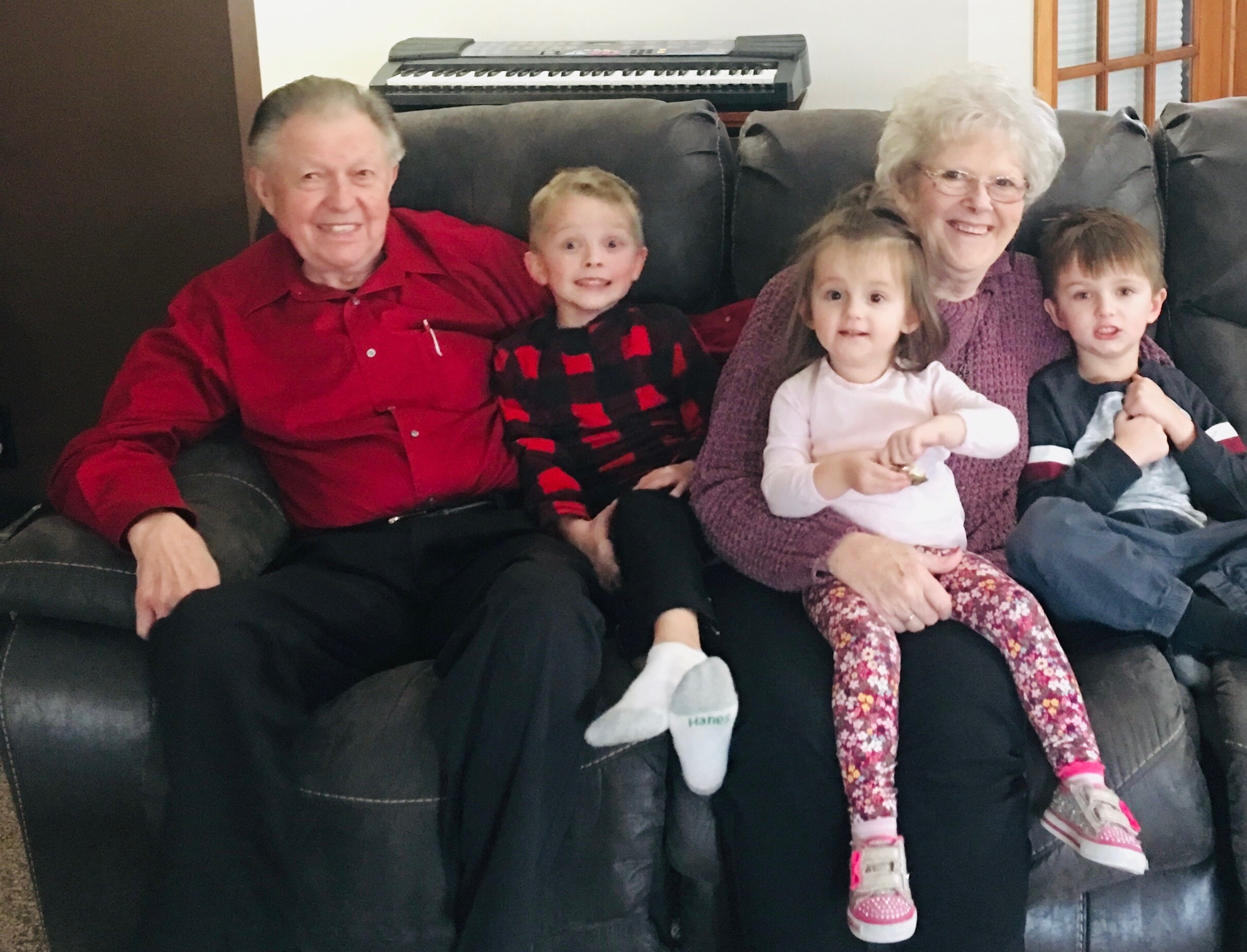 Smiling family of five seated on a dark leather couch: grandfather in red shirt, two young children, grandmother, and a boy on the right; keyboard in the background.