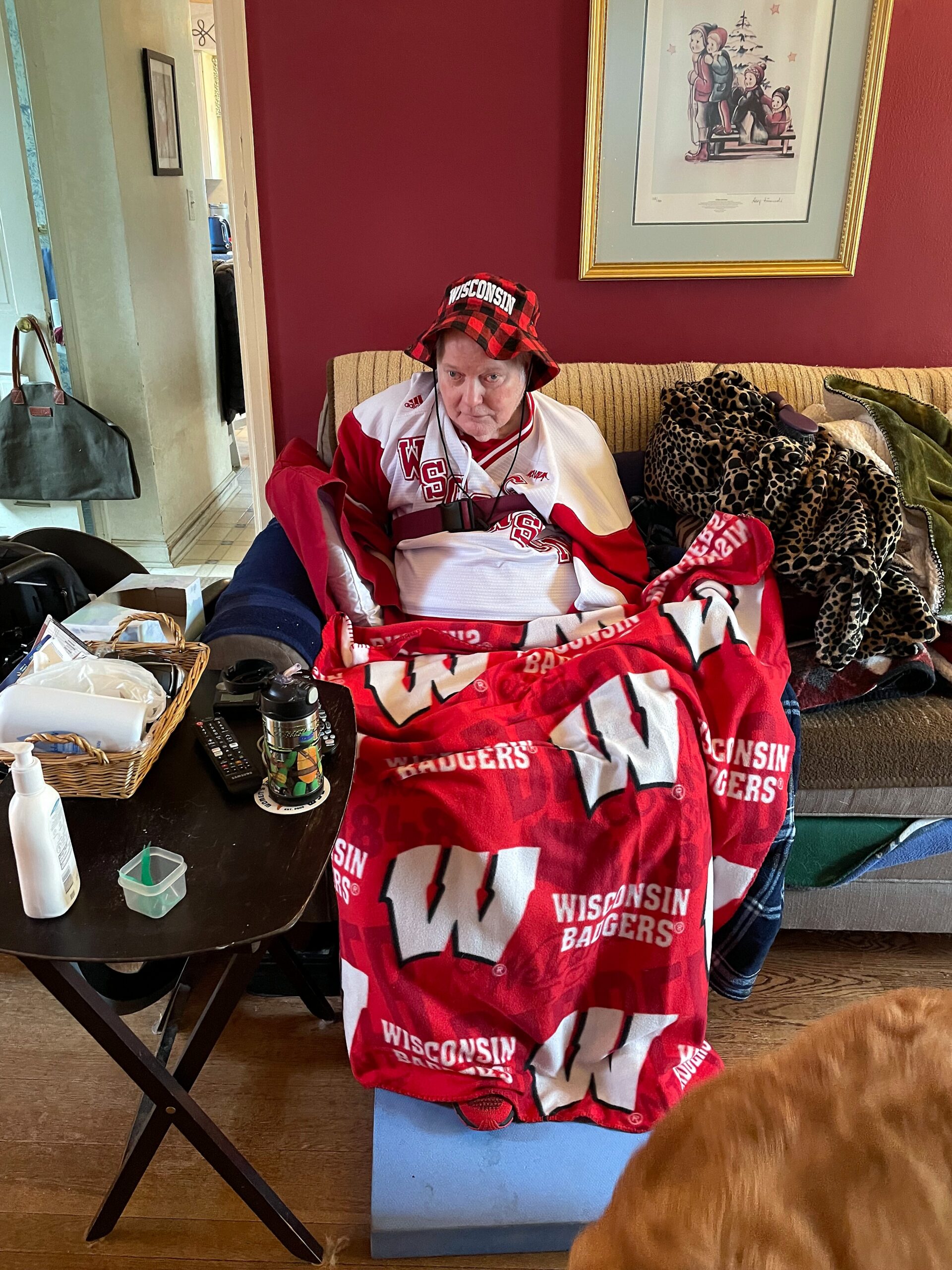 An older person wearing a Wisconsin Badgers hockey jersey and a red plaid bucket hat sits on a beige sofa, surrounded by blankets (leopard print and red Wisconsin), with a cluttered coffee table in front and a cat visible at the bottom edge of the image.
