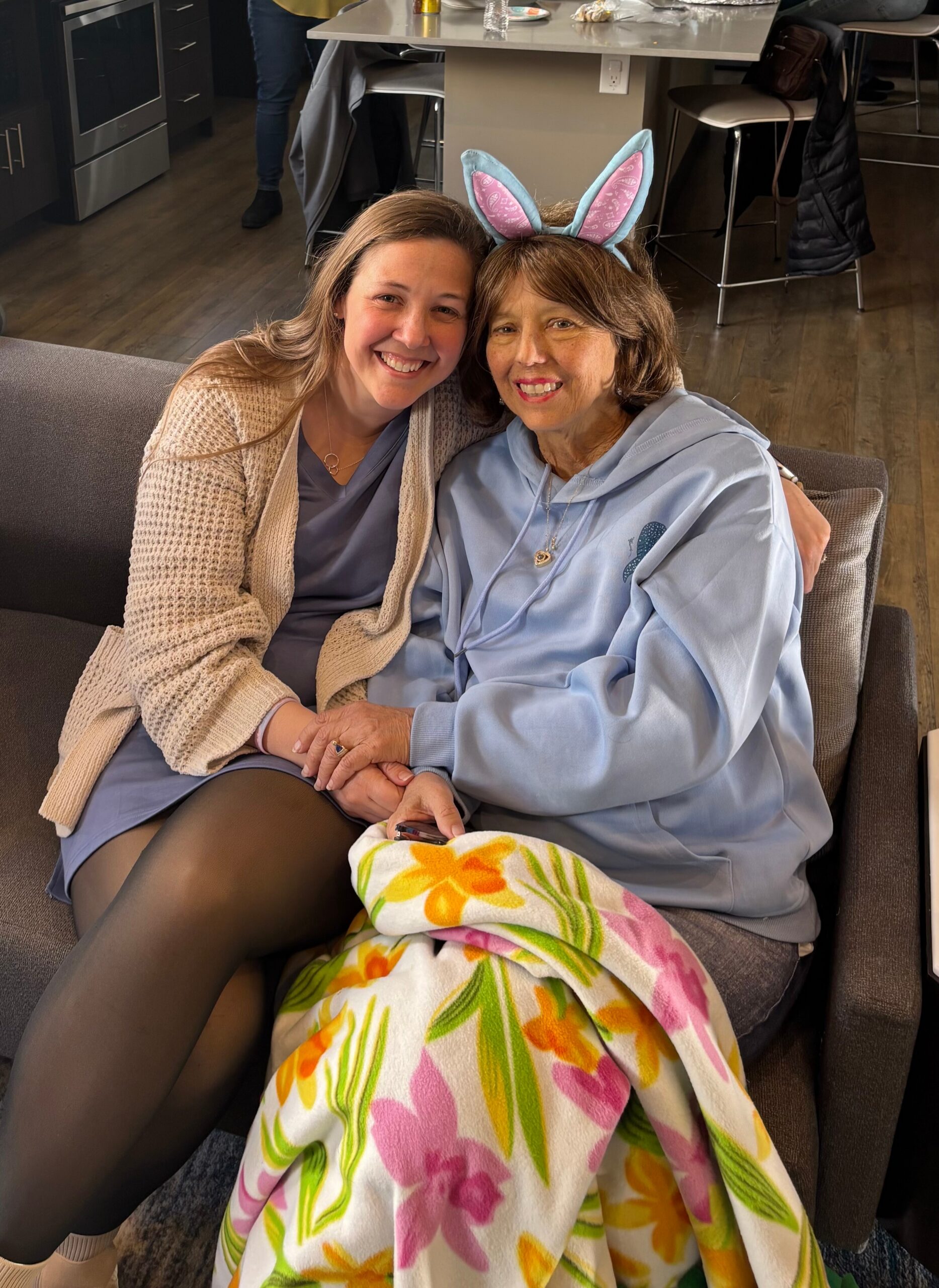 Two women sit close on a couch, smiling at the camera; one wears blue bunny ears and a pastel hoodie, the other a beige cardigan, both holding hands with a colorful floral blanket draped over their laps in a homey living room.