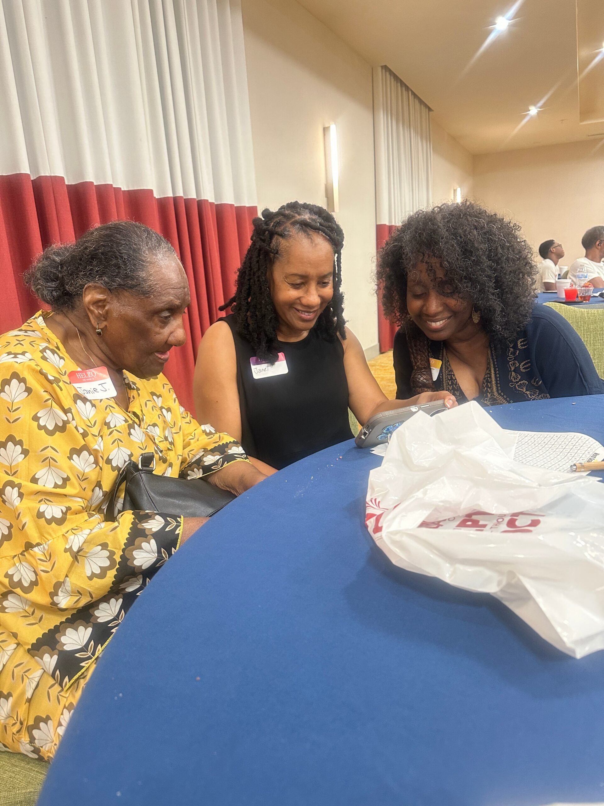 Three women sit at a blue table, smiling and looking at a shared device; left wears a yellow floral dress, middle in black, right in a dark patterned top, with name tags and a plastic bag on the table in a room with red-and-white curtains.