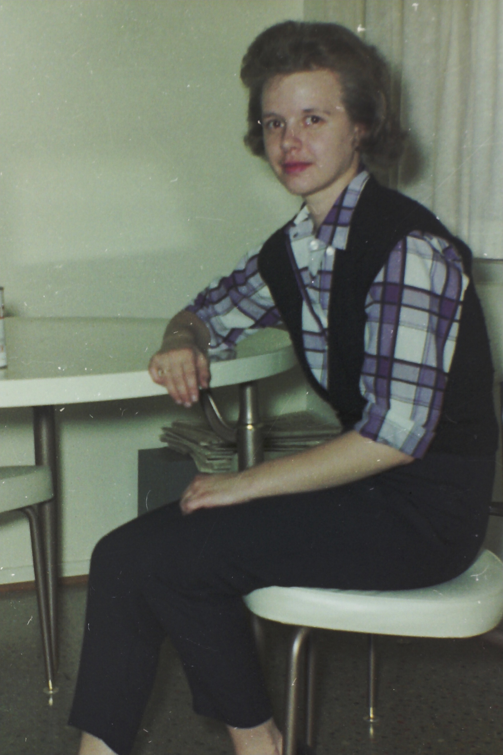 Woman sitting at a counter, wearing a purple plaid shirt and dark vest, looking at the camera in a home or office setting.