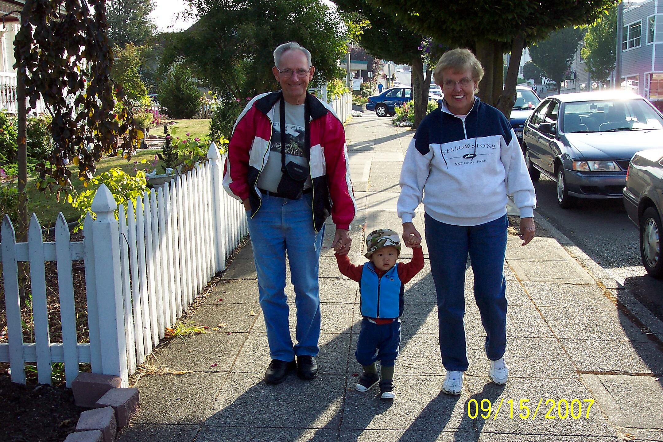 Family of three walking hand-in-hand on a sunny sidewalk beside a white picket fence in a suburban street.