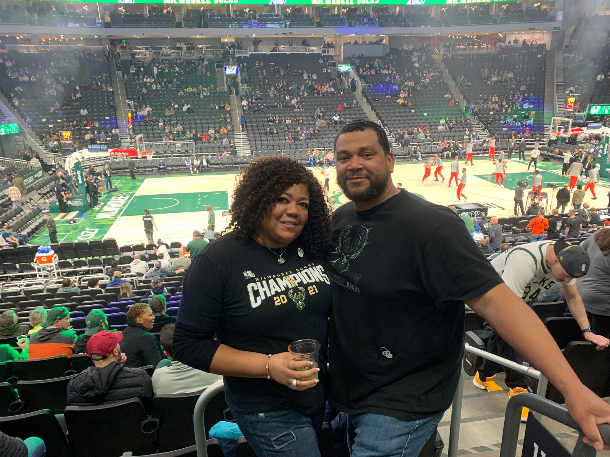 Two people smile for a selfie courtside at a basketball arena, with fans in the stands and players warming up on the court behind them, Bucks champions shirts visible.