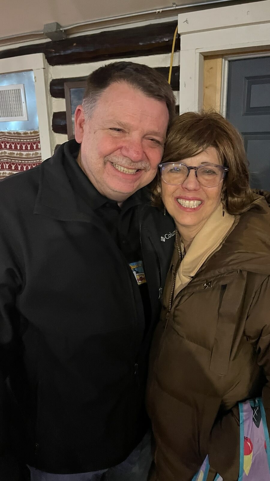 Two adults posing closely for a selfie, both smiling warmly inside a cozy room. (Man in black jacket left, woman in brown coat right)