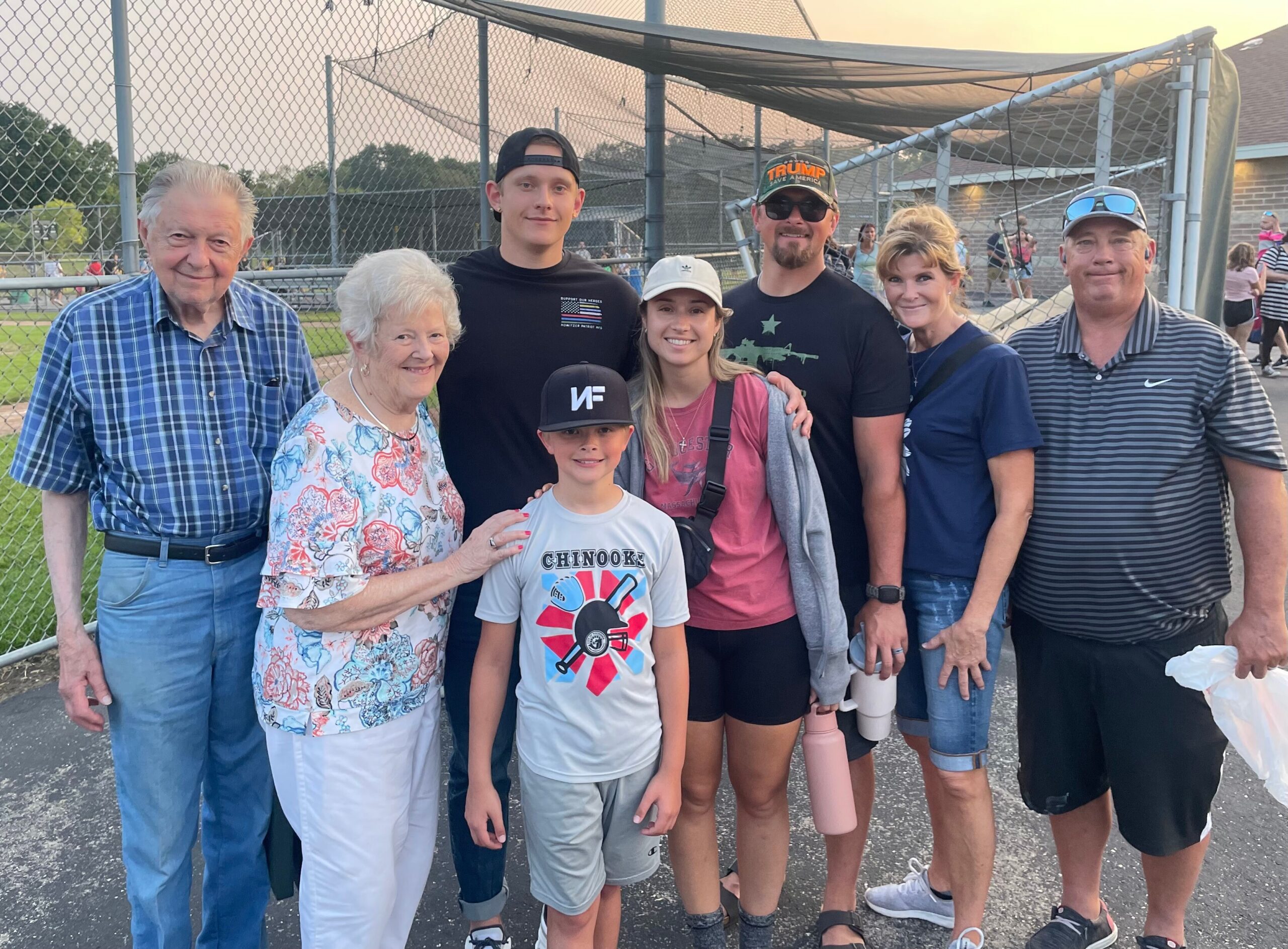 Group of people posing for a photo at a baseball field behind a chain-link fence, smiling at the camera.