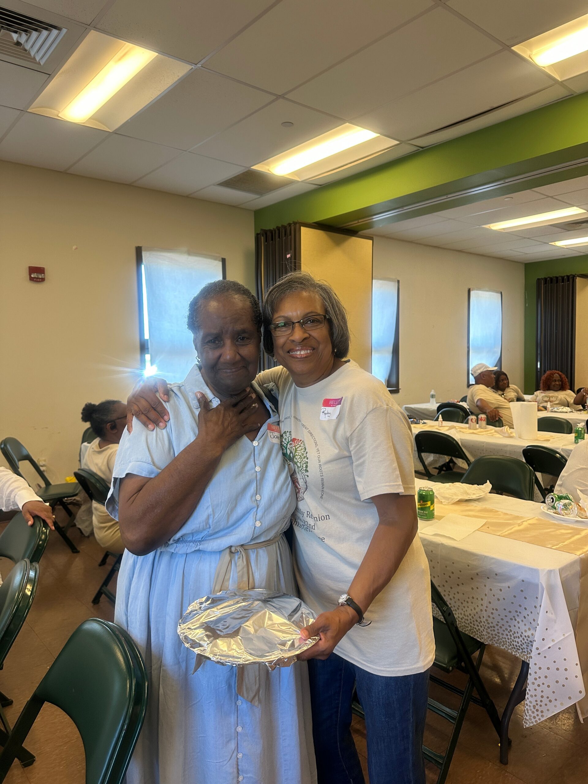 Two smiling women embrace at a community gathering, with tables set for a meal in the background.