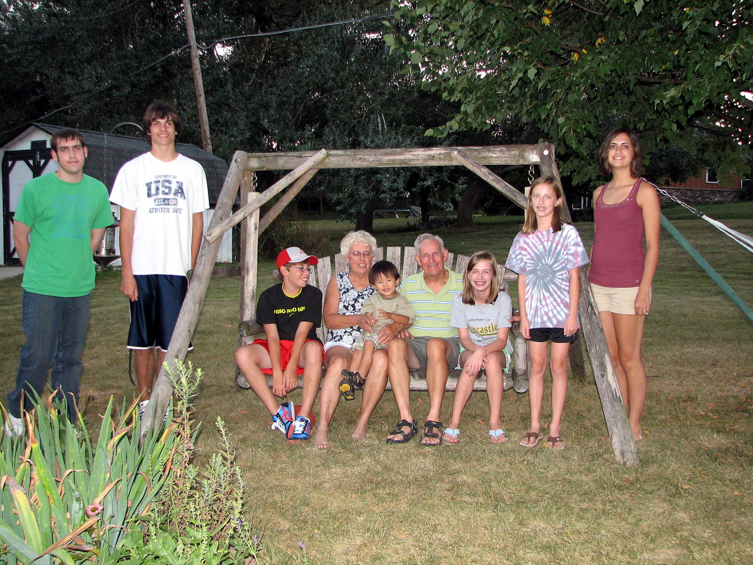 Group of ten people, including children and adults, posing on and around a wooden swing frame in a backyard park setting with plants in the foreground.