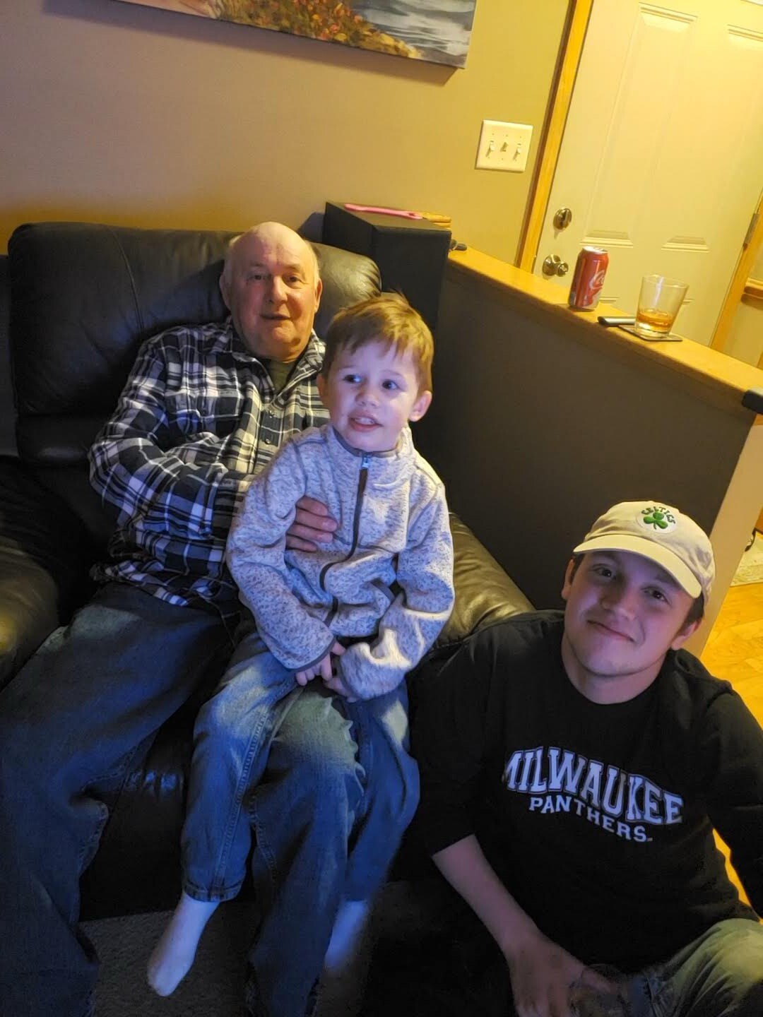 Three generations together in a living room: an elderly man on a black leather couch holds a smiling toddler on his lap, while a young man in a Milwaukee Panthers shirt sits beside them and wears a cap.