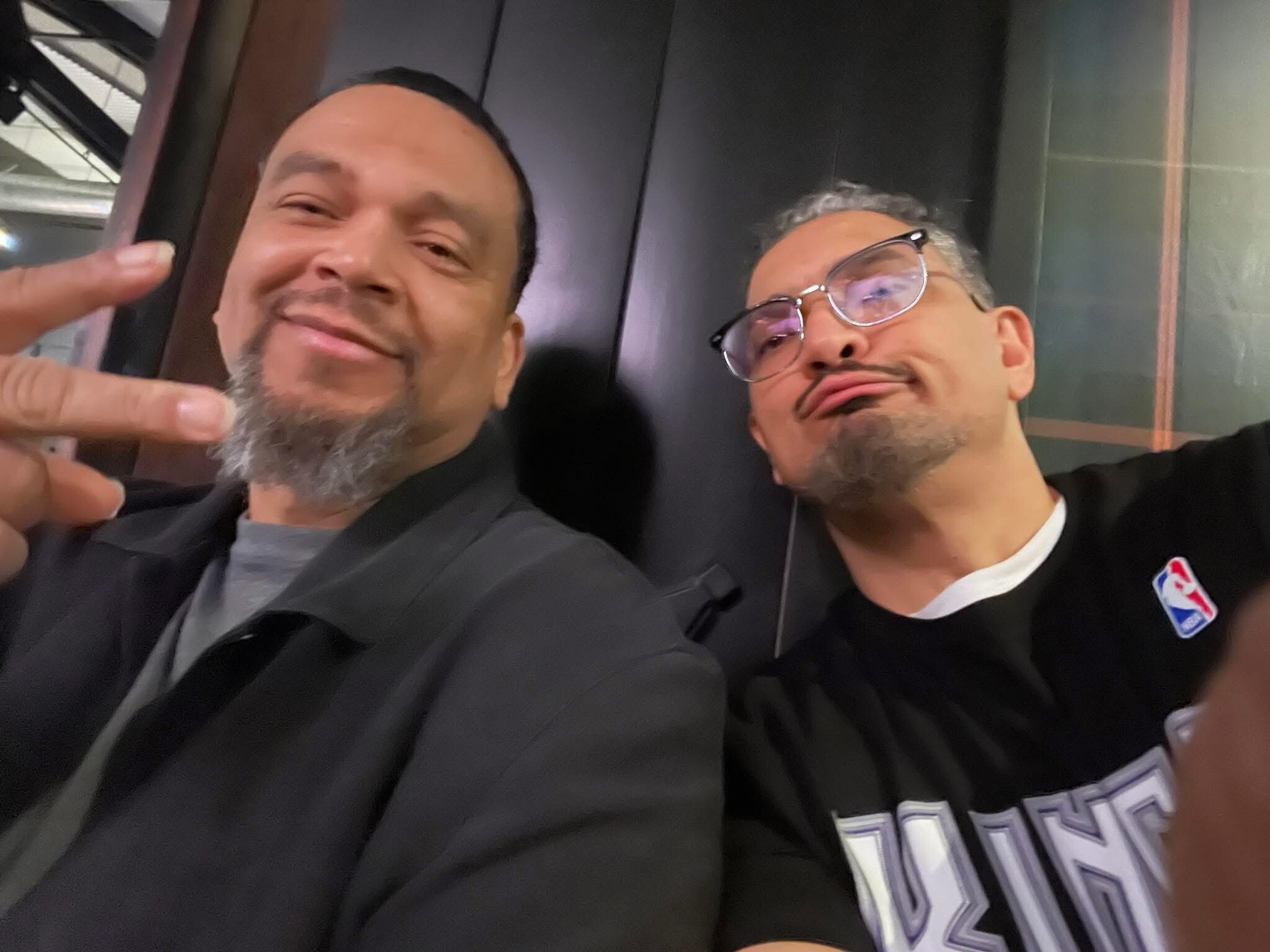 Two men taking a close-up selfie indoors, wearing dark jerseys; one man points at the camera with a peace sign while smiling.