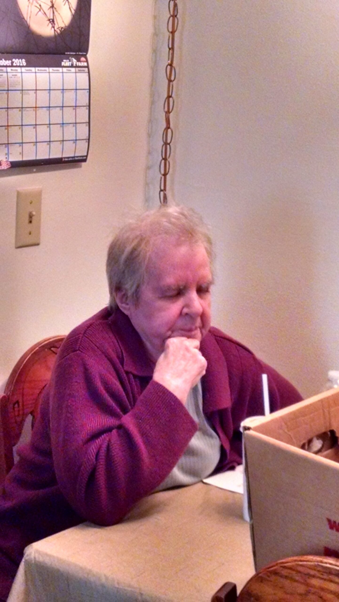 An older woman with short gray hair sits at a table with her hand under her chin, eyes closed, wearing a maroon cardigan in a dining area with a calendar on the wall behind her.