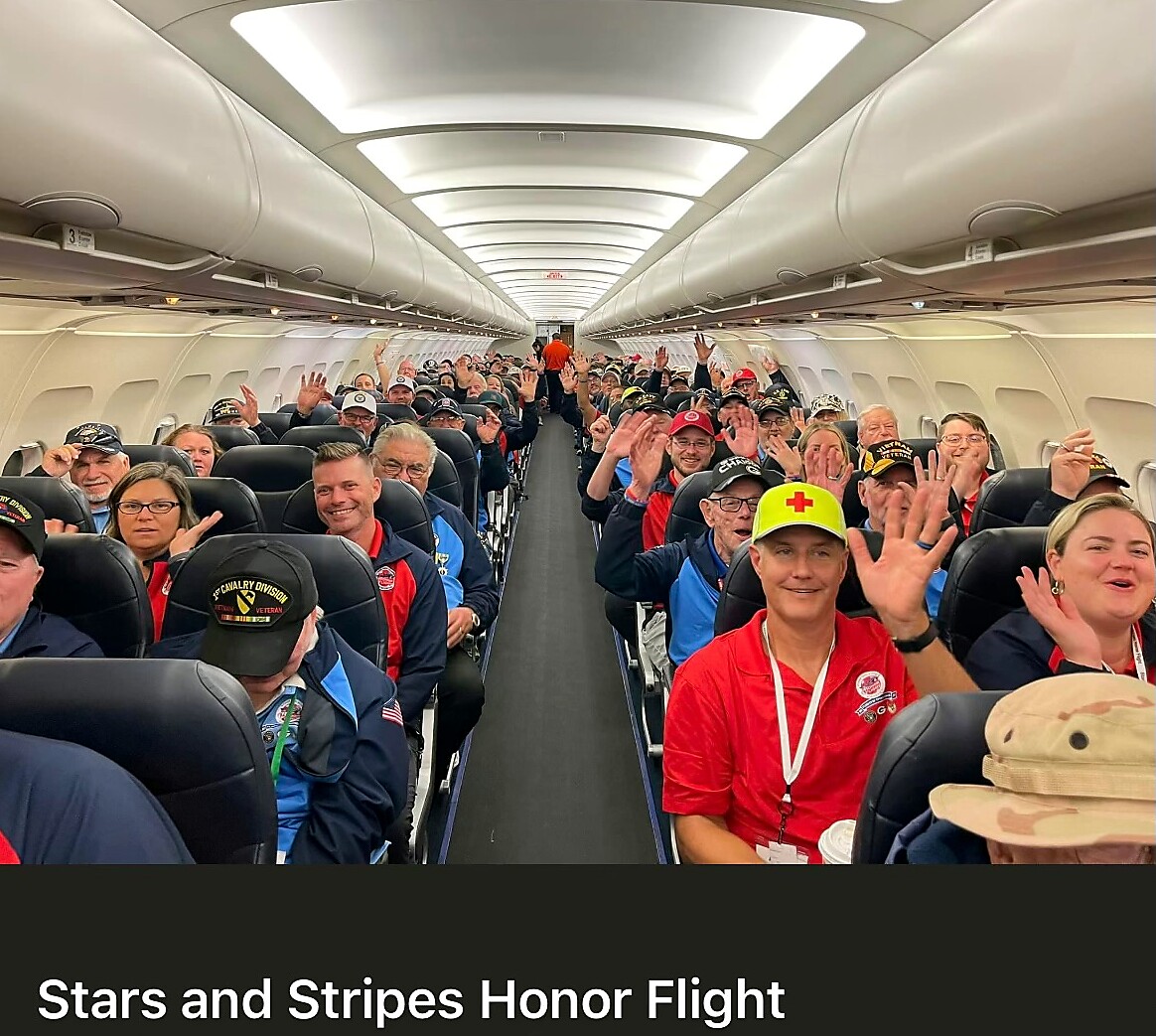 A large group of veterans and volunteers sit in an airplane cabin, many waving at the camera during a Stars and Stripes Honor Flight.