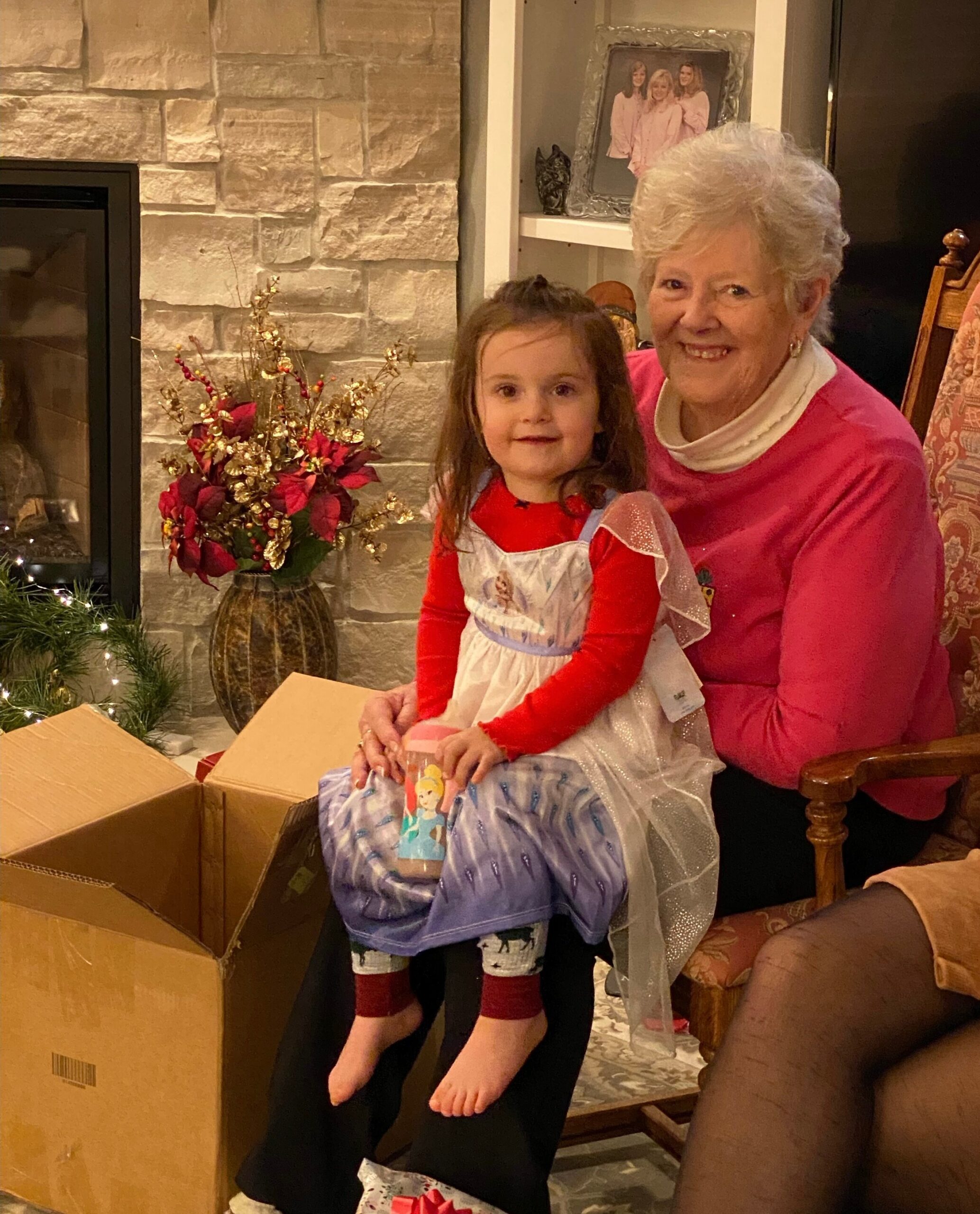 A smiling grandmother and young girl sit together on a couch with gift boxes and holiday decorations nearby in a cozy living room.