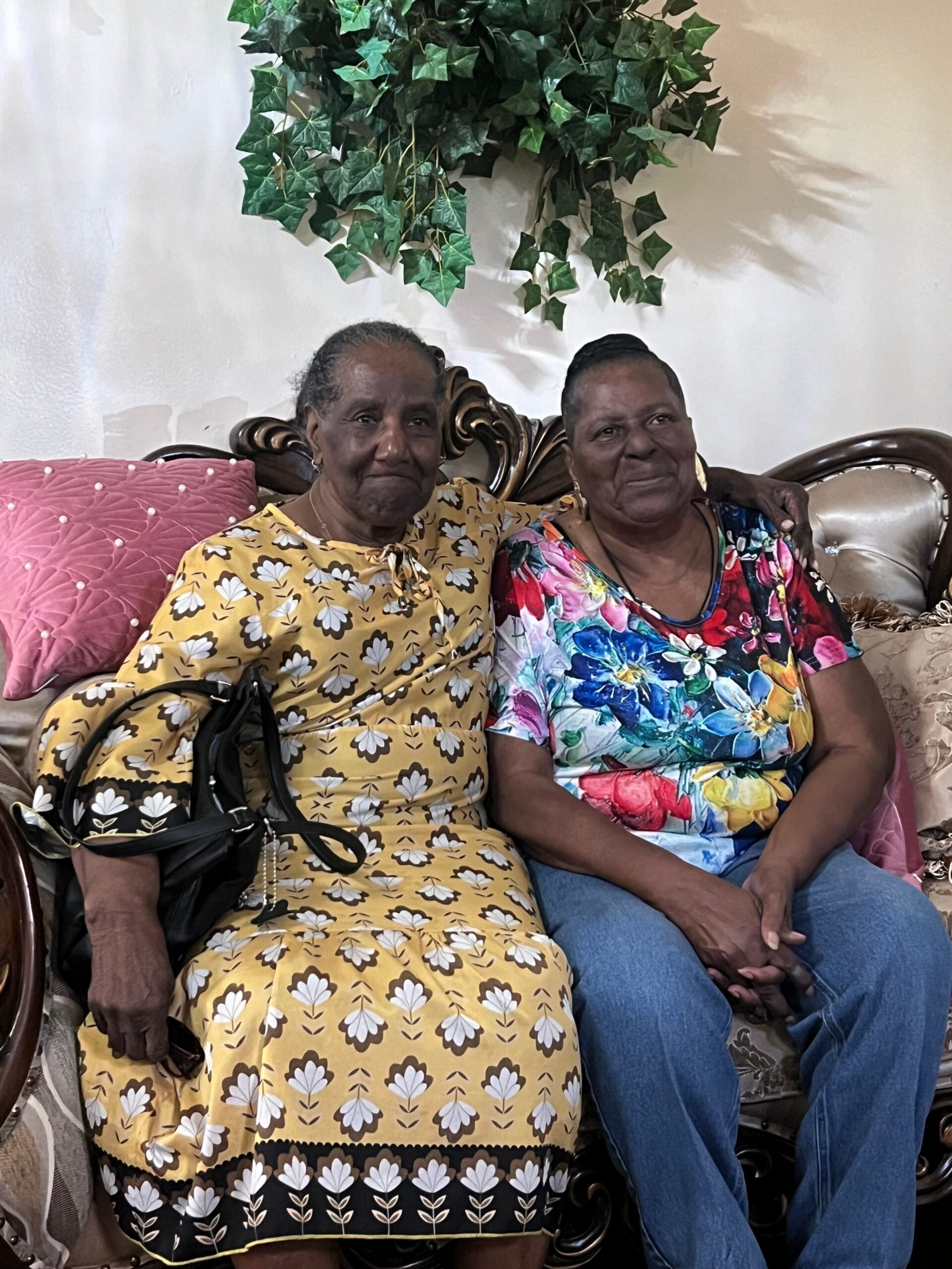 Two elderly women sit close together on a carved sofa; one wears a yellow floral dress, the other a bright multicolored top with a leafy plant overhead.