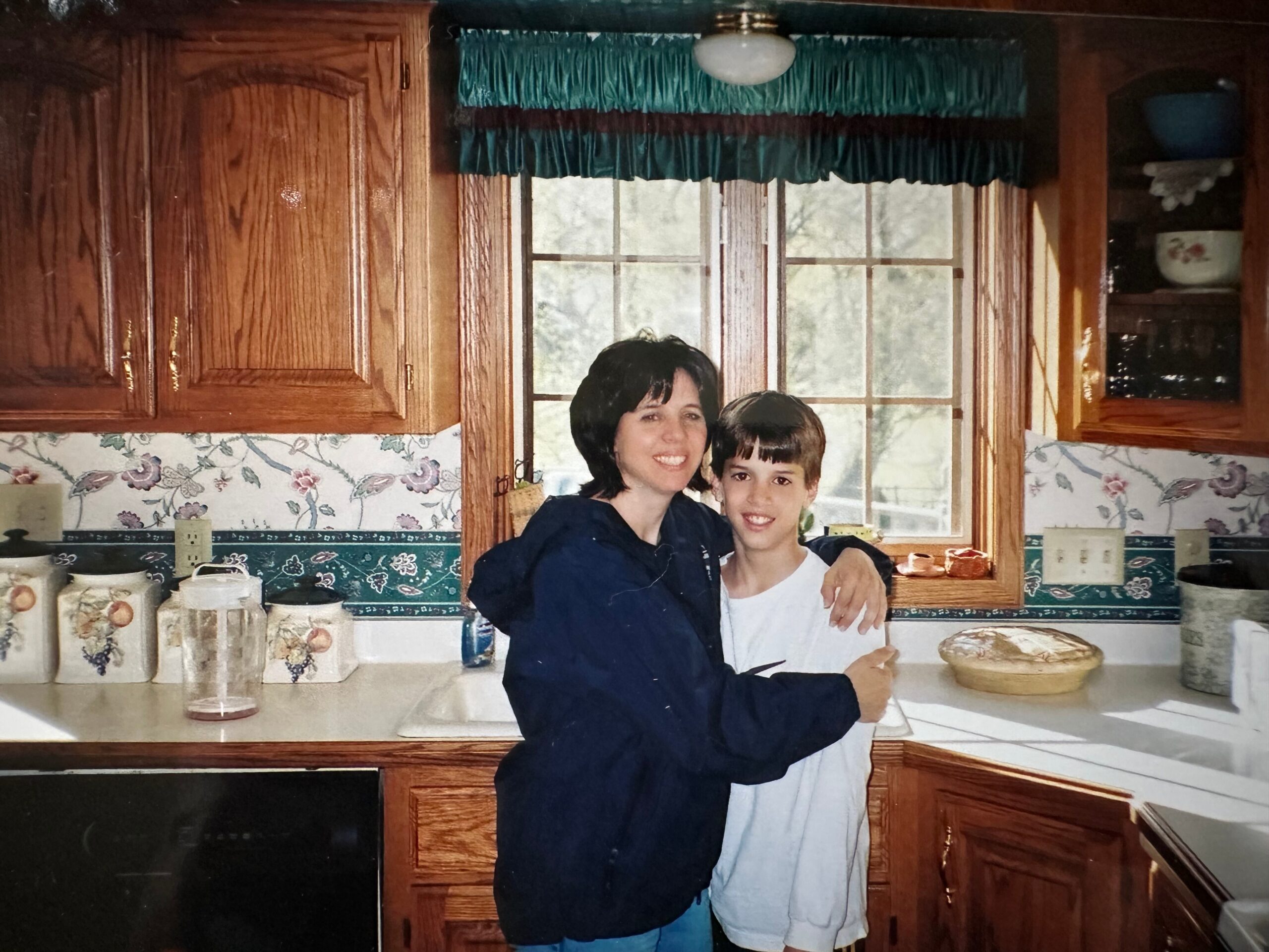 Smiling woman and child pose together in a warm wooden kitchen, with a window behind them and a pie on the counter.