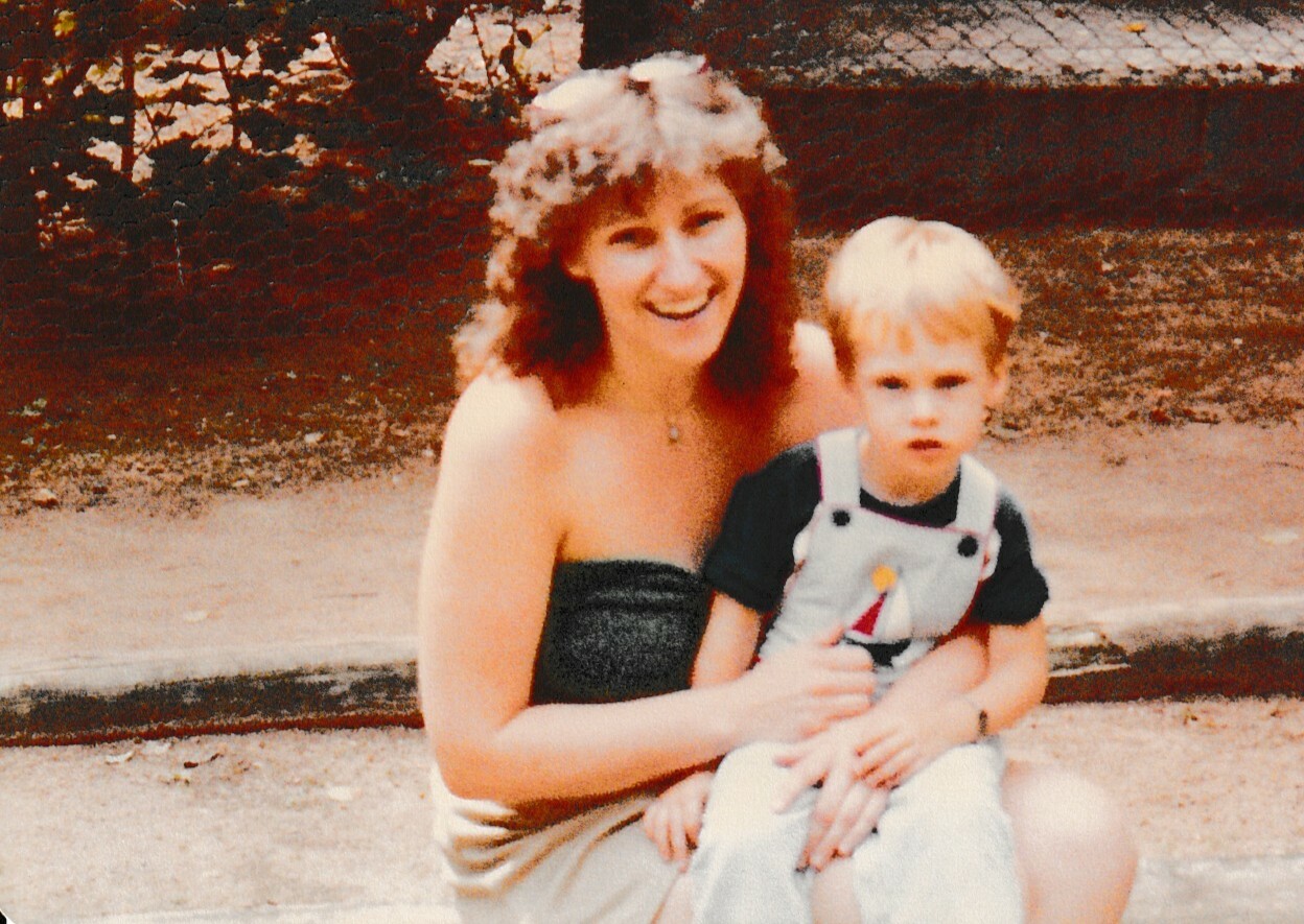 Smiling woman with curly hair holding a young boy in overalls outdoors on a sunny day