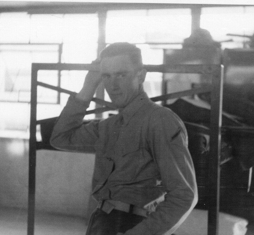 Black-and-white photo of a man in a light shirt leaning on a metal railing, one hand resting on his head, indoors with large windows in the background.