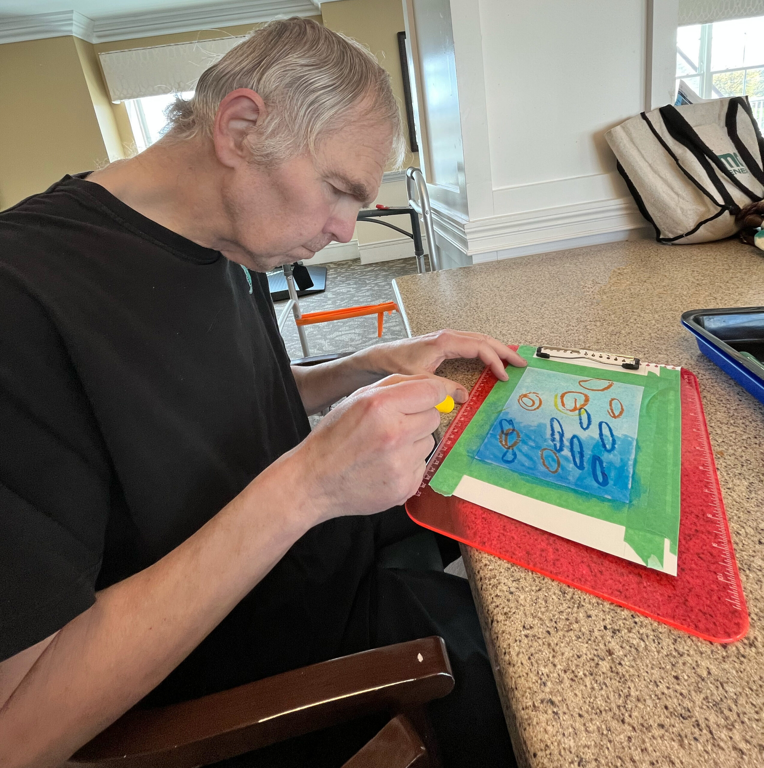 Man with gray hair wearing a black shirt sits at a counter, working with colored paper, a ruler, and small craft pieces on a red cutting mat.