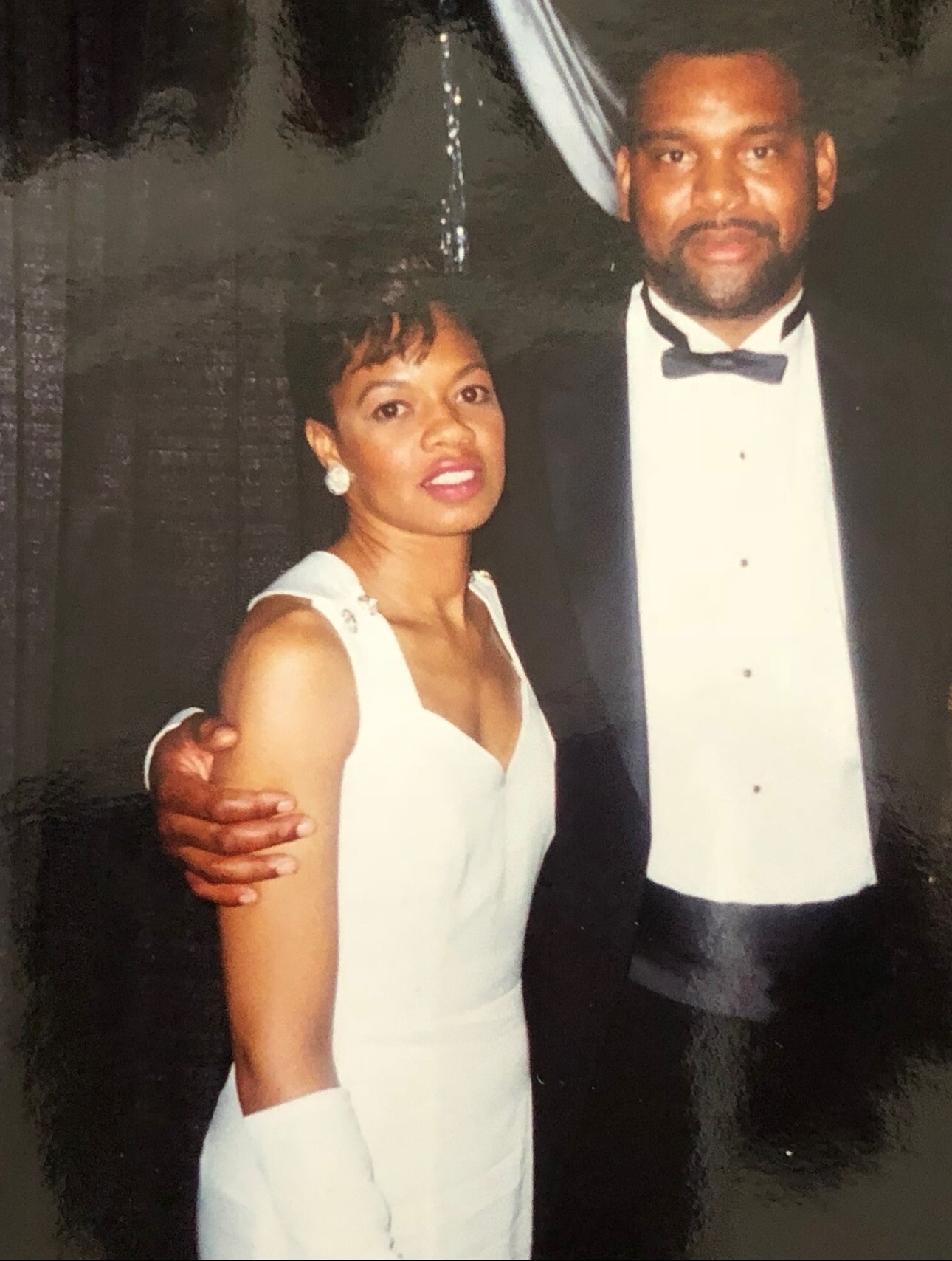A couple dressed formally posing together; woman in a white gown with earrings, man in a black tuxedo and bow tie, with arms around each other.