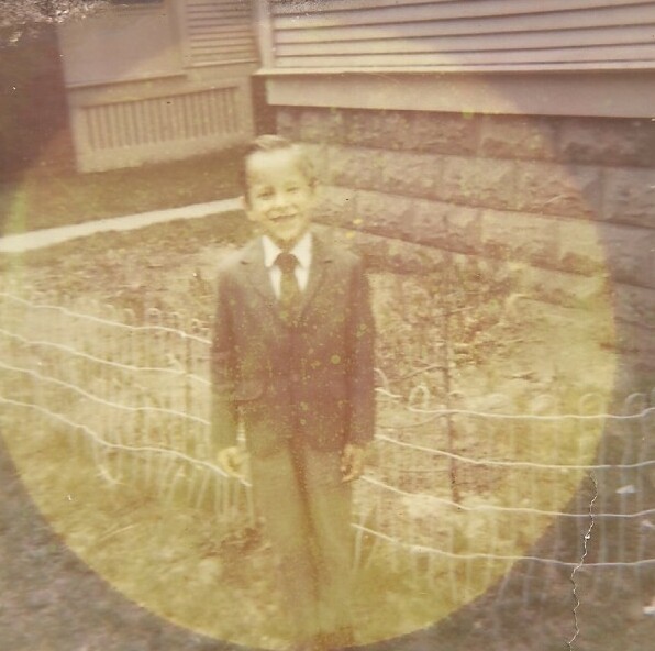 Boy in a suit and tie standing outside a house with a stone foundation and a wire fence.