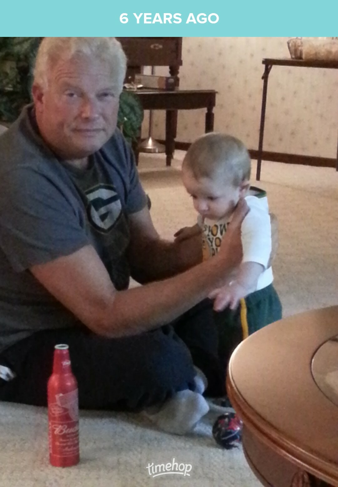 Older man with gray hair sits on a carpet, holding a toddler who stands beside him in a living room setting.