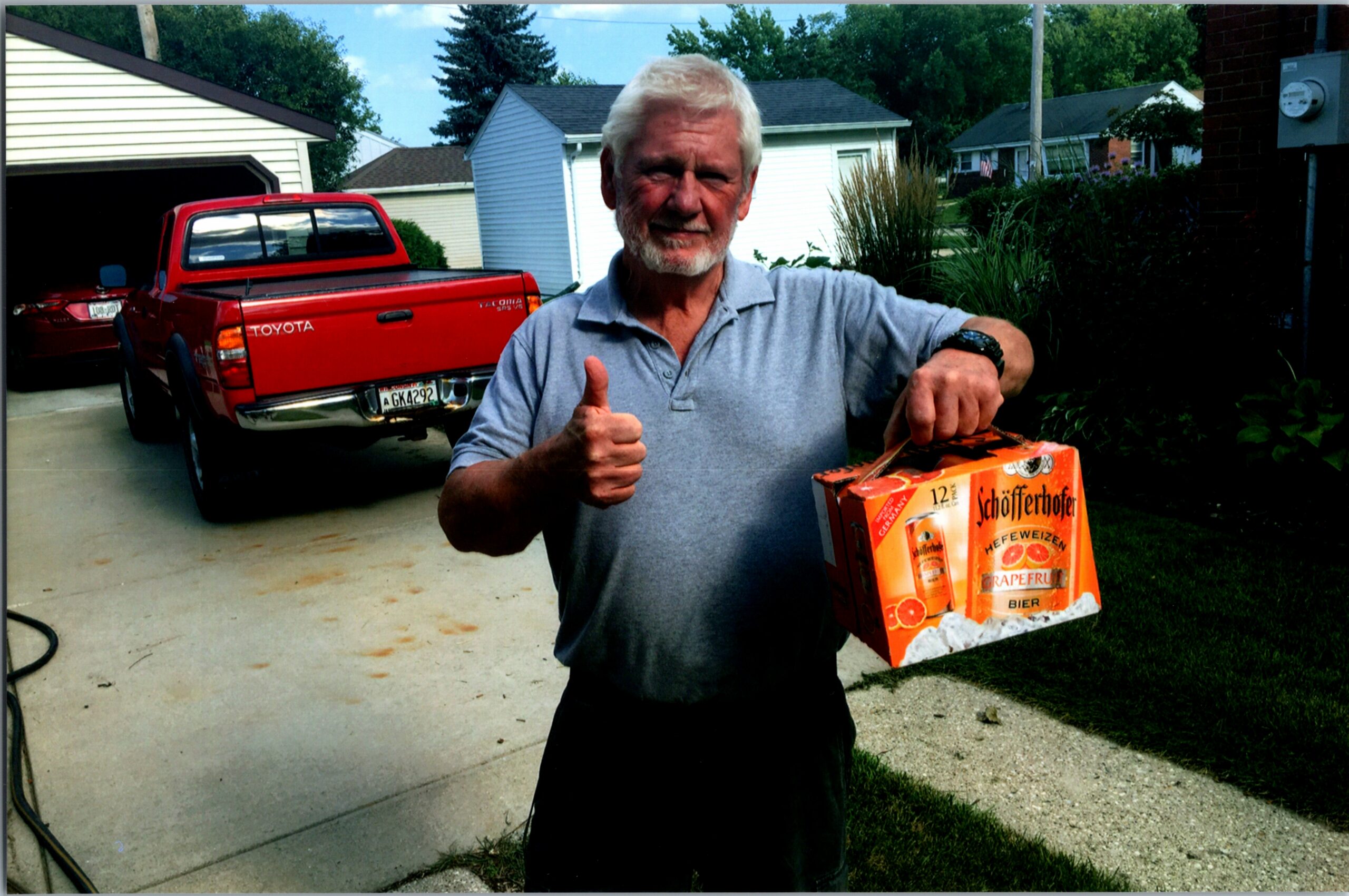 Older man with white hair and beard giving a thumbs-up while holding a box of Schöfferhofer grapefruit beer in a driveway with a red Toyota truck behind him
