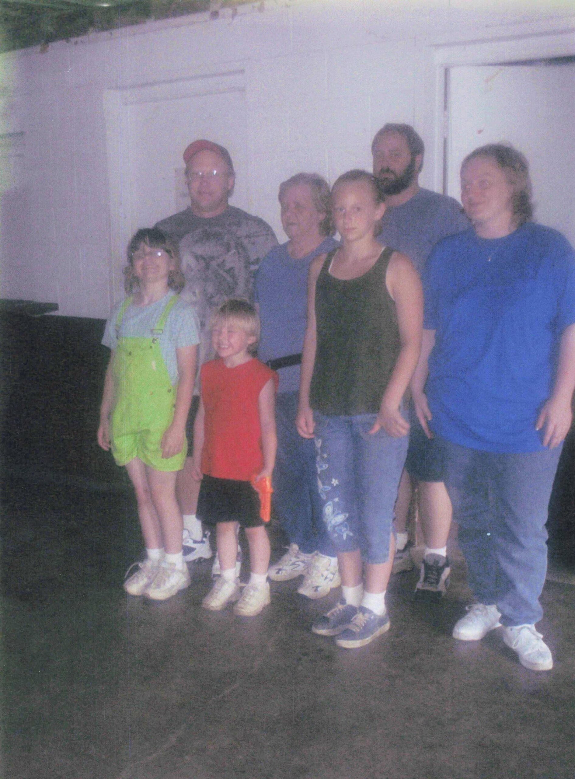 Group of seven people posing indoors, including two children in front (girl in lime-green overalls and boy in red shirt) and adults behind them in casual clothing.