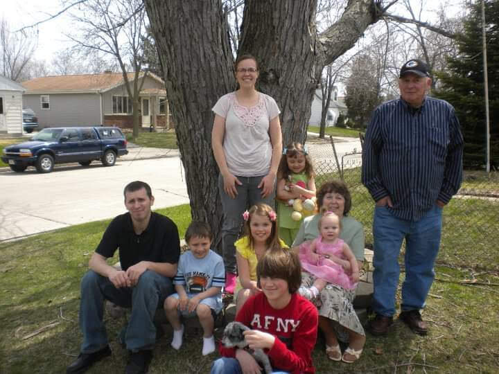 Family portrait outdoors under a large tree in a suburban yard, with adults and children smiling and posing together.