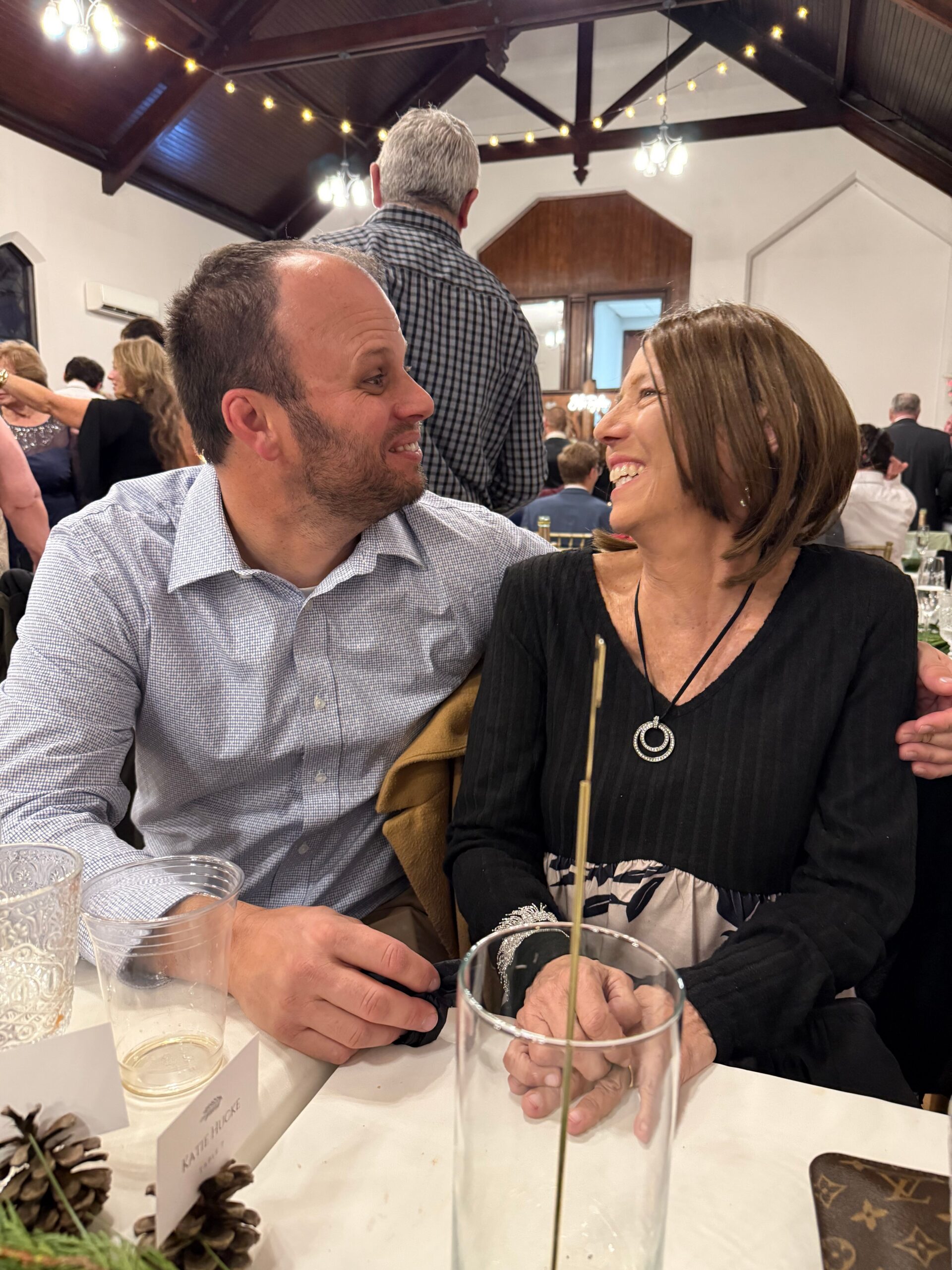 A smiling man and woman sit side by side at a festive table, looking at each other warmly during a crowded indoor event, with drinks in front of them.