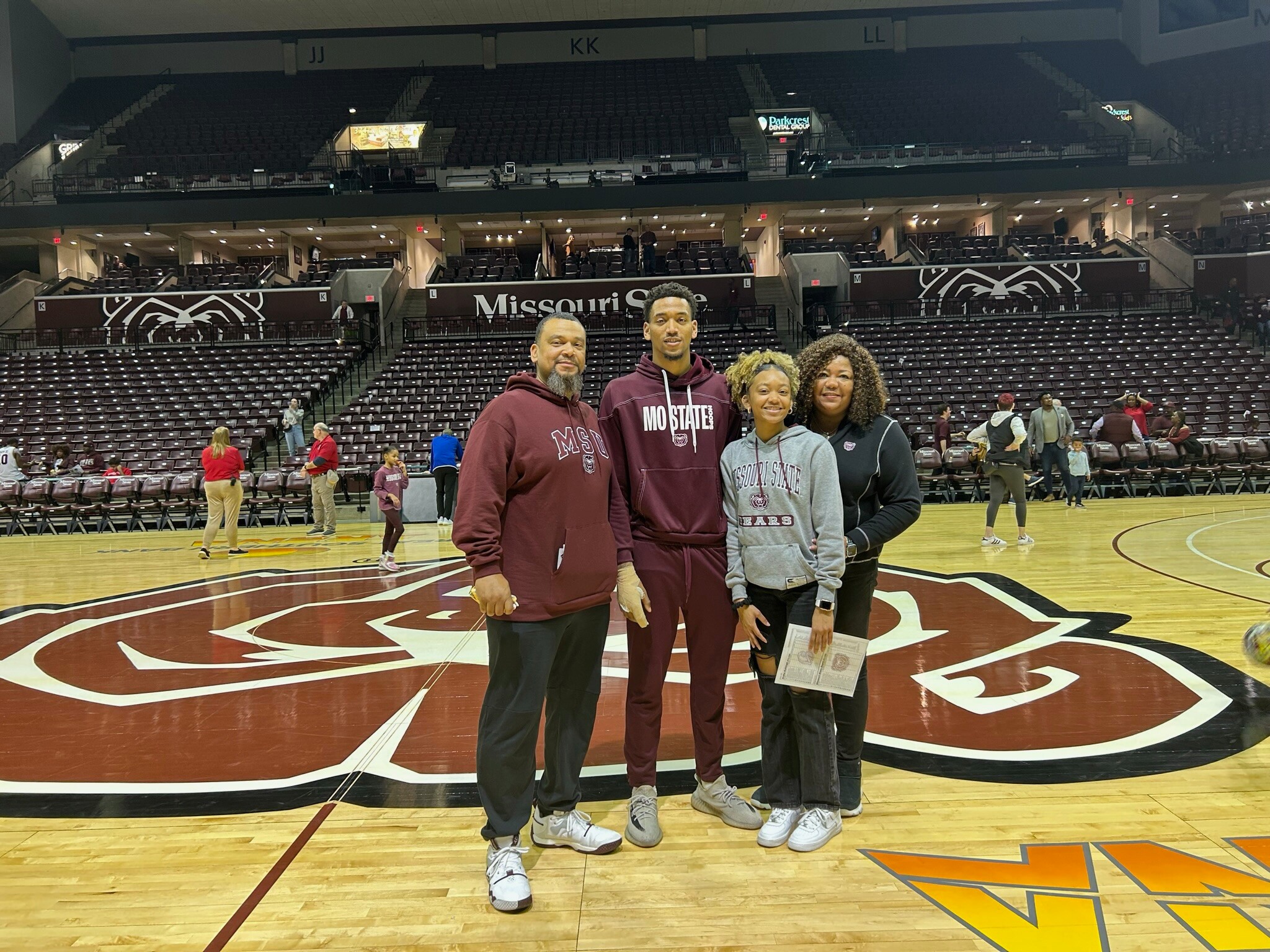 Group of four people posing on a basketball court in an arena, wearing MSU gear and smiling at the camera with empty stands behind them.