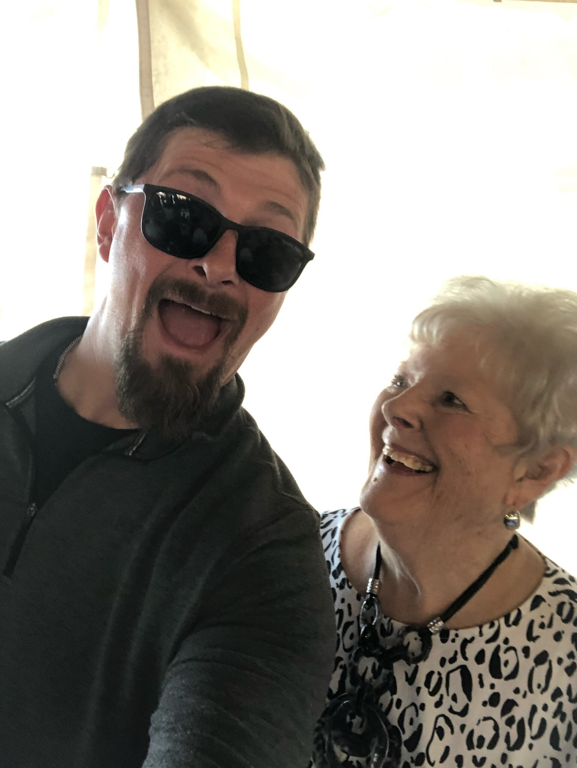 Two people taking a selfie: man with sunglasses and goatee on the left, smiling older woman on the right in a leopard-print blouse.