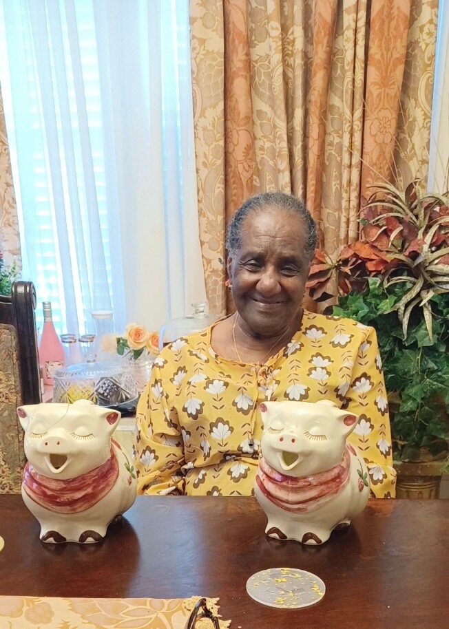 Smiling elderly woman sits at a table with two white piggy bank figurines wearing red scarves beside her in a patterned room setting.