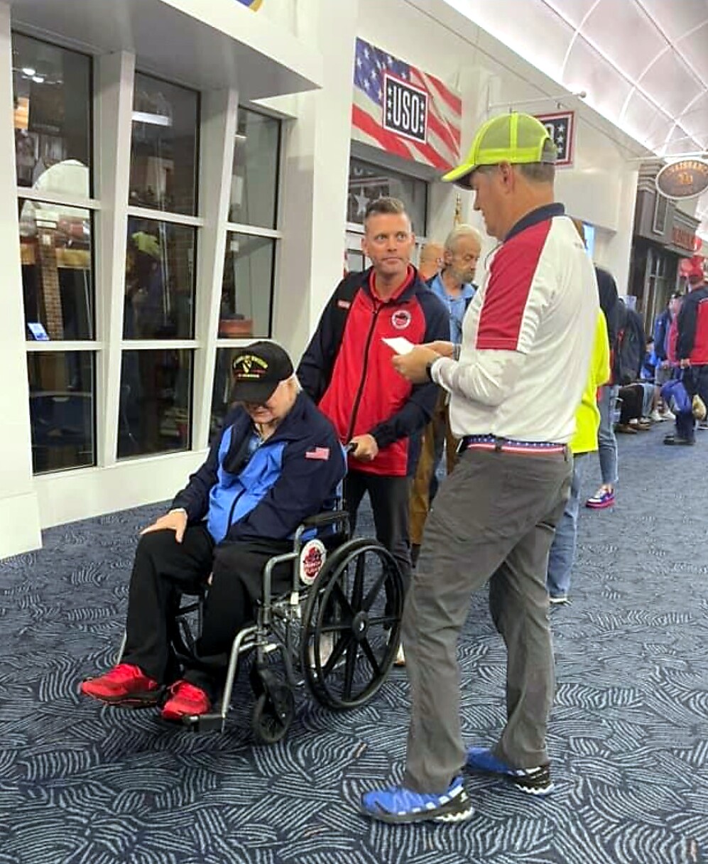 An elderly man in a wheelchair being assisted by a man in a red-and-black jacket in a busy indoor venue with USO flag in the background.