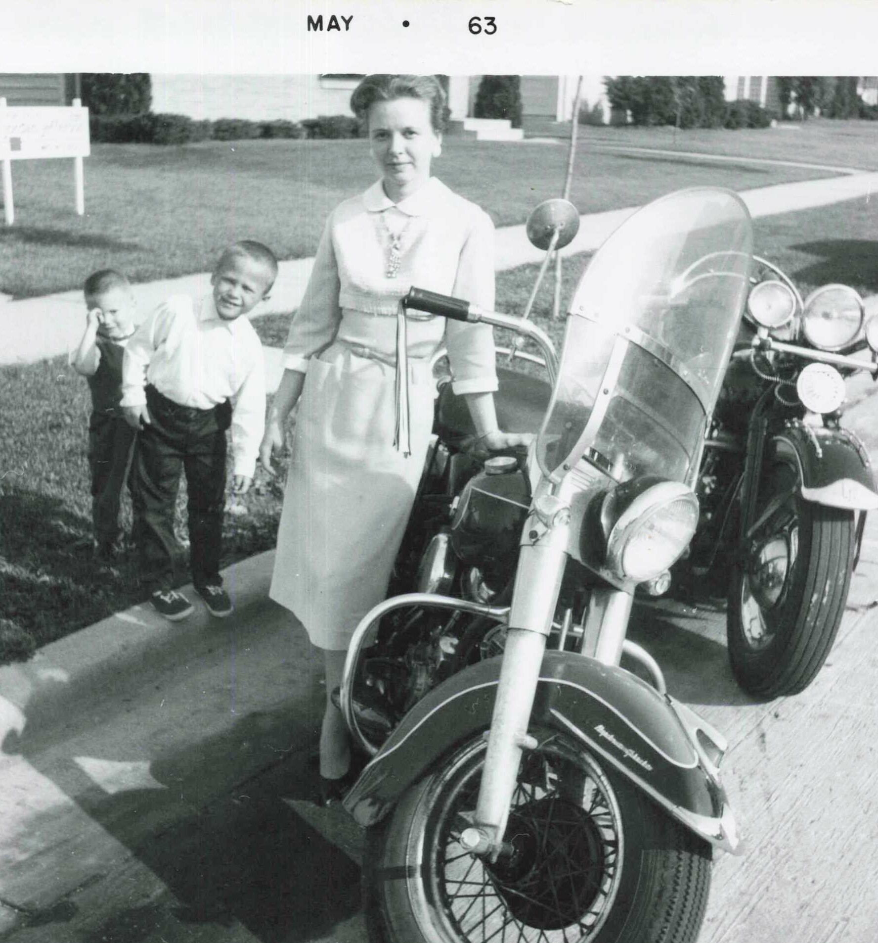 A woman in a light dress stands beside a large motorcycle with two young boys nearby in a suburban yard.